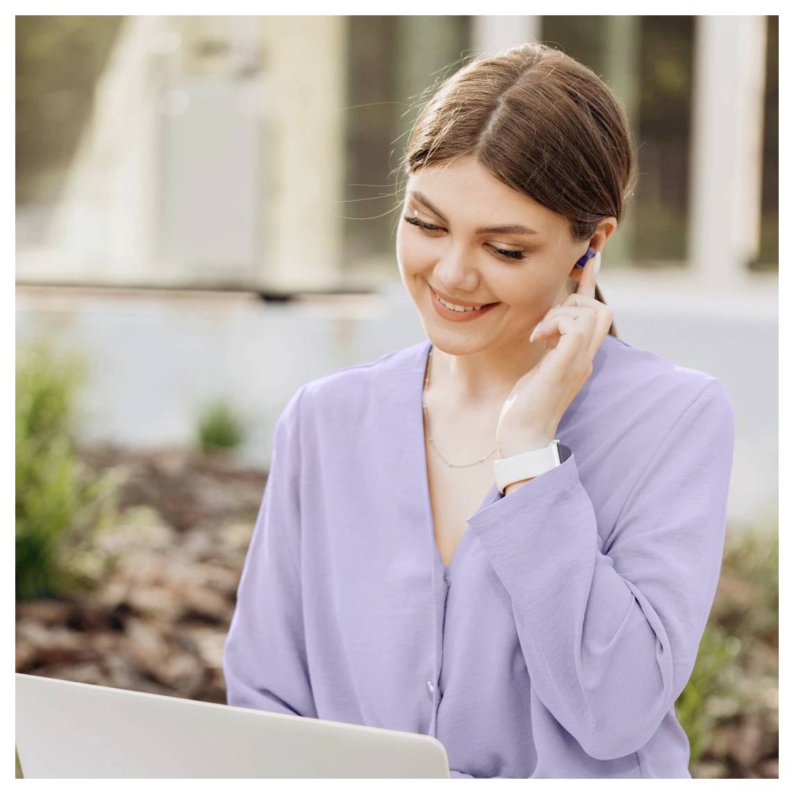 Lächelnde Frau in einer lavendelfarben Bluse stellt ein Ohrhörer ein, während sie auf einen Laptop im Freien schaut.