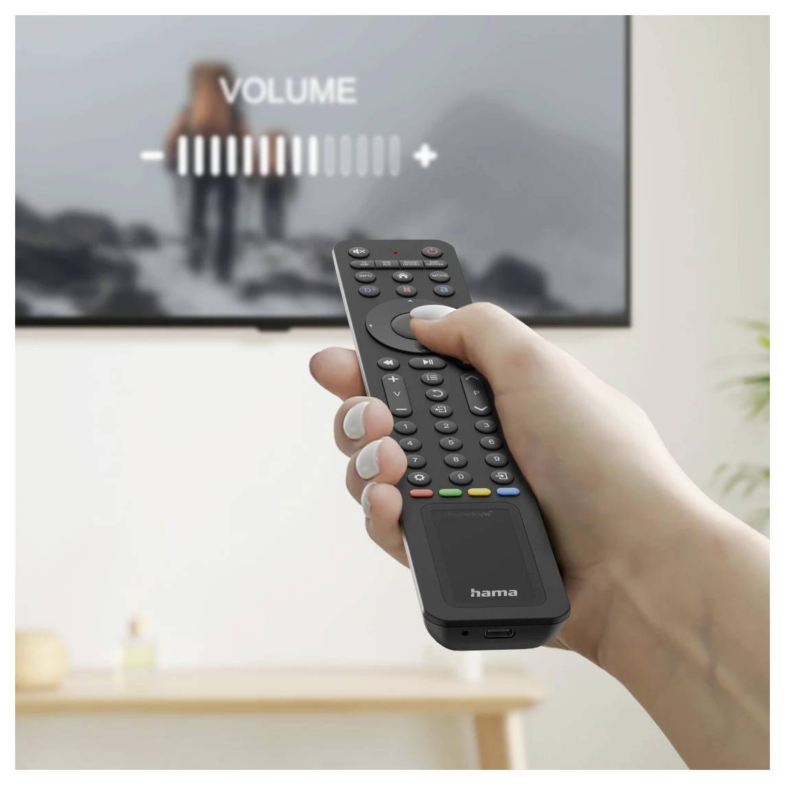 Hand hält eine Fernbedienung, während die Lautstärke auf dem Bildschirm angepasst wird, mit einem unscharfen Fernseher, der 