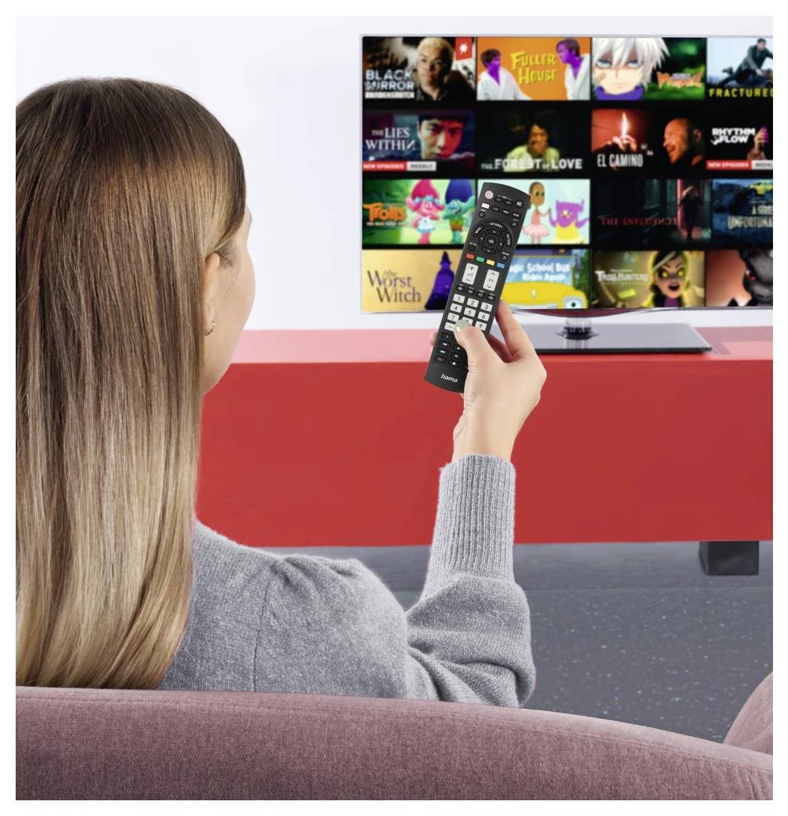 Person auf einem Sofa, das eine Fernbedienung hält und durch ein Streaming-App-Menü auf einem Fernsehbildschirm mit Show- und Film-Miniaturansichten blättert.