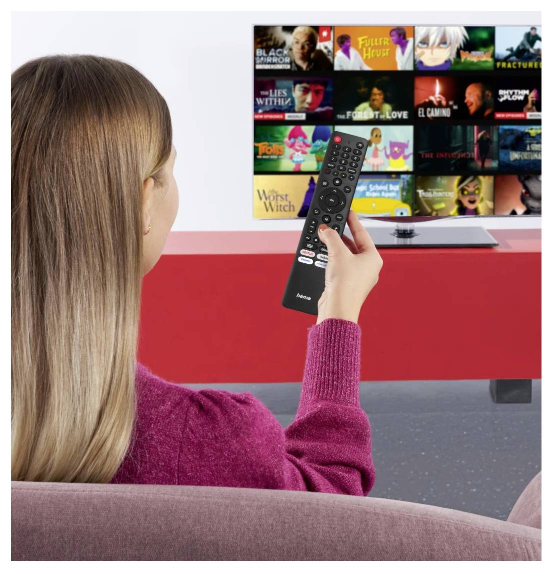 Frau auf einem Sofa zeigt mit einer Fernbedienung auf einen Smart-TV, der ein Raster mit Miniaturansichten von Streaming-Filmen und -Serien anzeigt.