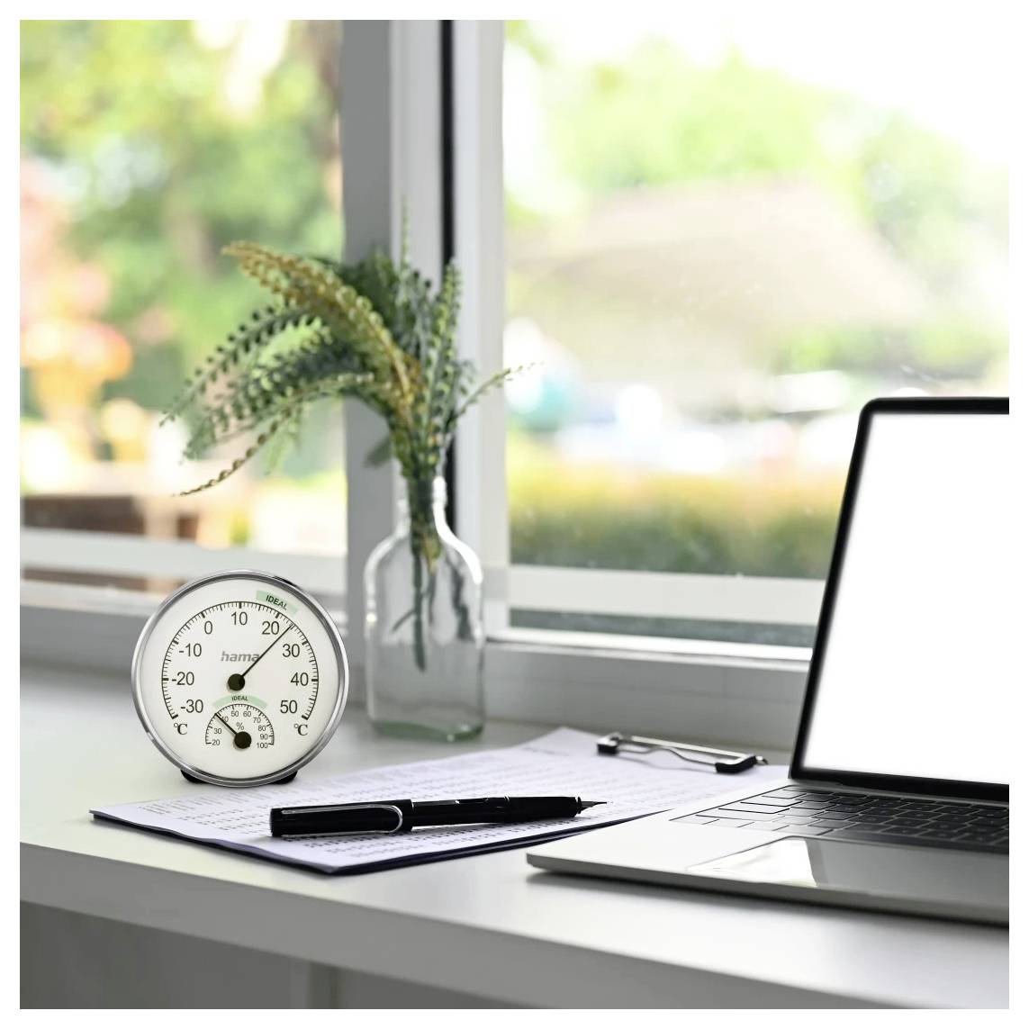Laptop auf einem Schreibtisch neben einem Fenster mit einem Blatt Papier, einem Stift, einer Klemmbrett, einem Thermometer und einer Vase mit Blumen in einem hellen Homeoffice.