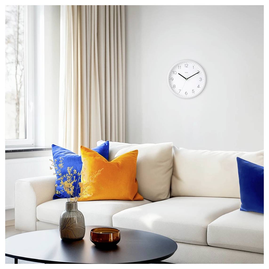 Helles modernes Wohnzimmer mit weißem Sofa, blauen und orangefarbenen Kissen, runder Wanduhr und schwarzem Couchtisch neben einem Fenster.