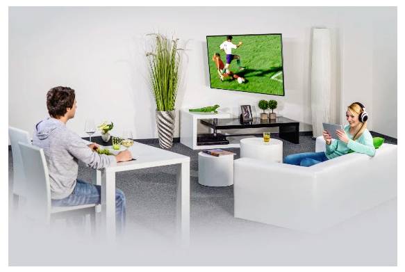 Zwei Menschen entspannen sich in einem Wohnzimmer und schauen sich ein Fußballspiel auf einem an der Wand montierten Fernseher an, wobei einer an einem Tisch sitzt und der andere auf einem Sofa liegt und ein Tablet nutzt.