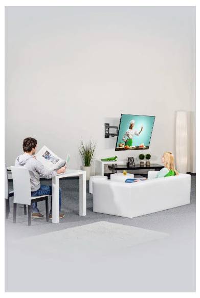 Ein Mann liest eine Zeitschrift an einem Tisch, während ein Kind auf einem Sofa sitzt und im hellen Wohnzimmer Fernsehen schaut.
