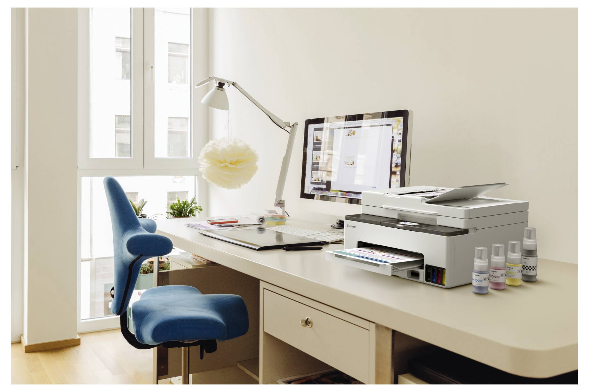 Helles Homeoffice mit blauem Stuhl, Schreibtisch, Computer, Drucker, Lampe und Zubehör neben einem Fenster.