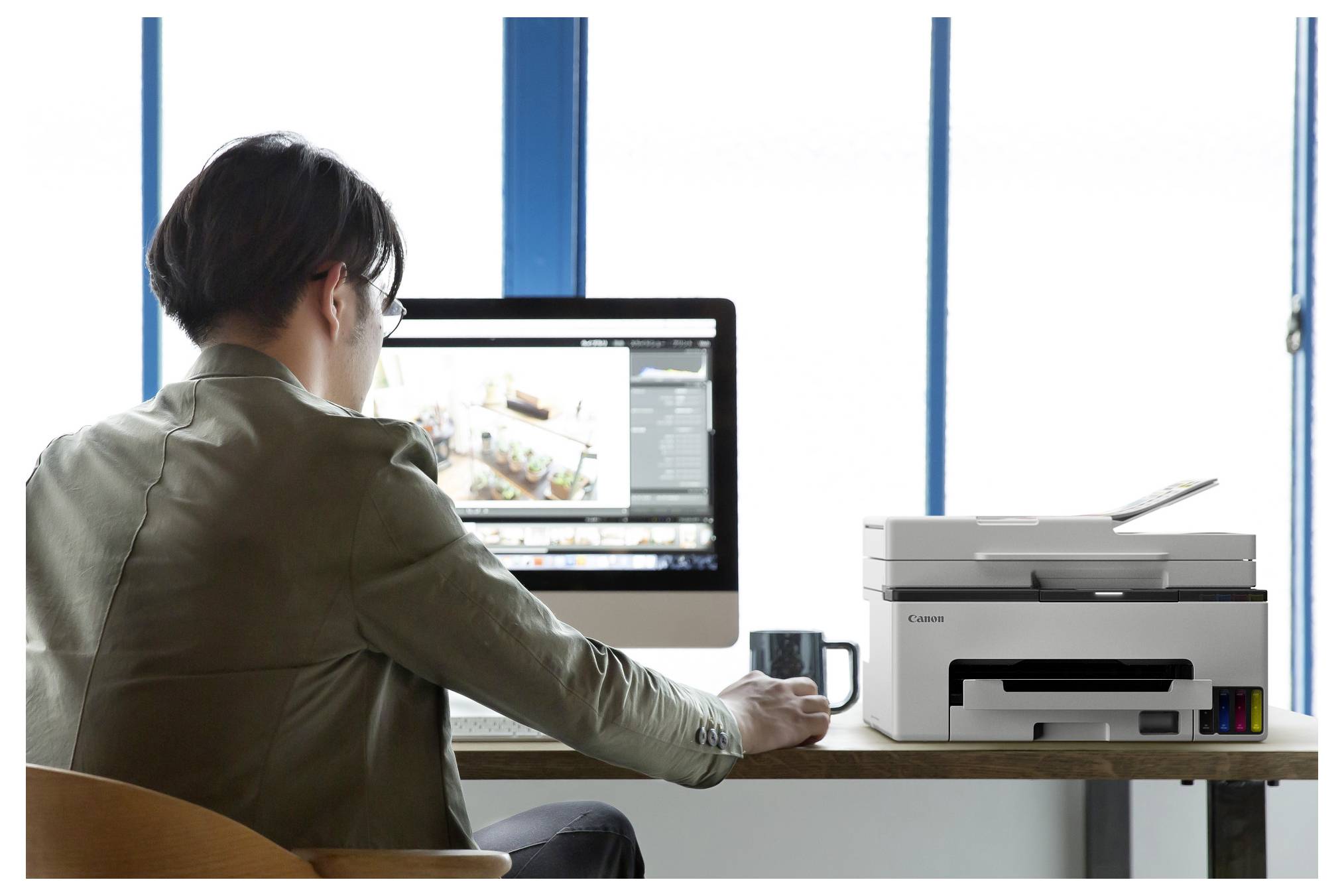 Eine Person, die an einem Desktop-Computer neben einem Drucker in einem hellen Büro arbeitet, von hinten betrachtet.