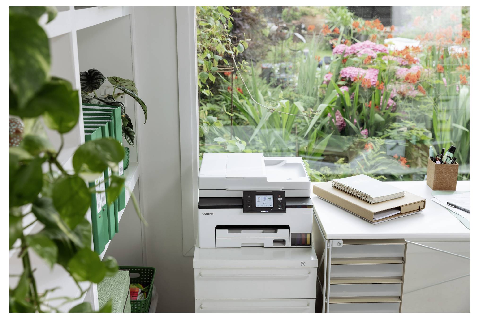 Homeoffice-Ecke mit einem Canon-Drucker neben einem Schreibtisch an einem Fenster mit Ausblick auf einen üppigen Garten mit rosa Blüten.