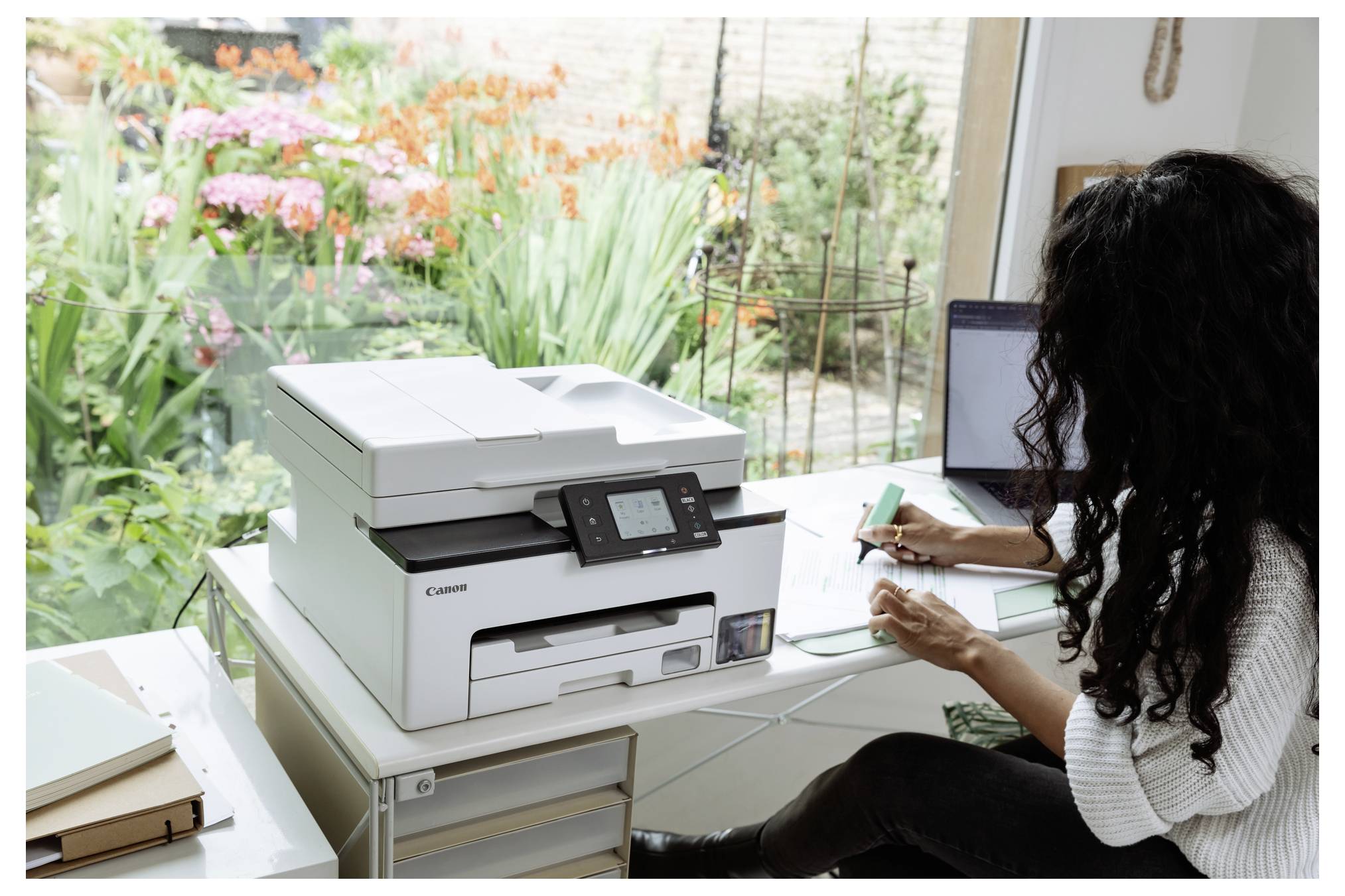 Frau arbeitet an einem Schreibtisch neben einem Canon-Drucker und schreibt auf Papier in einem Heimbüro mit einem Laptop und Blick auf den Garten.