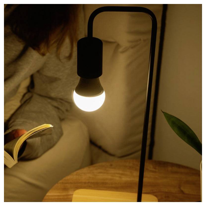 Eine Person liest ein Buch bei einer kleinen leuchtenden Lampe auf einem Nachttisch in einem gemütlichen, schwach beleuchteten Zimmer.