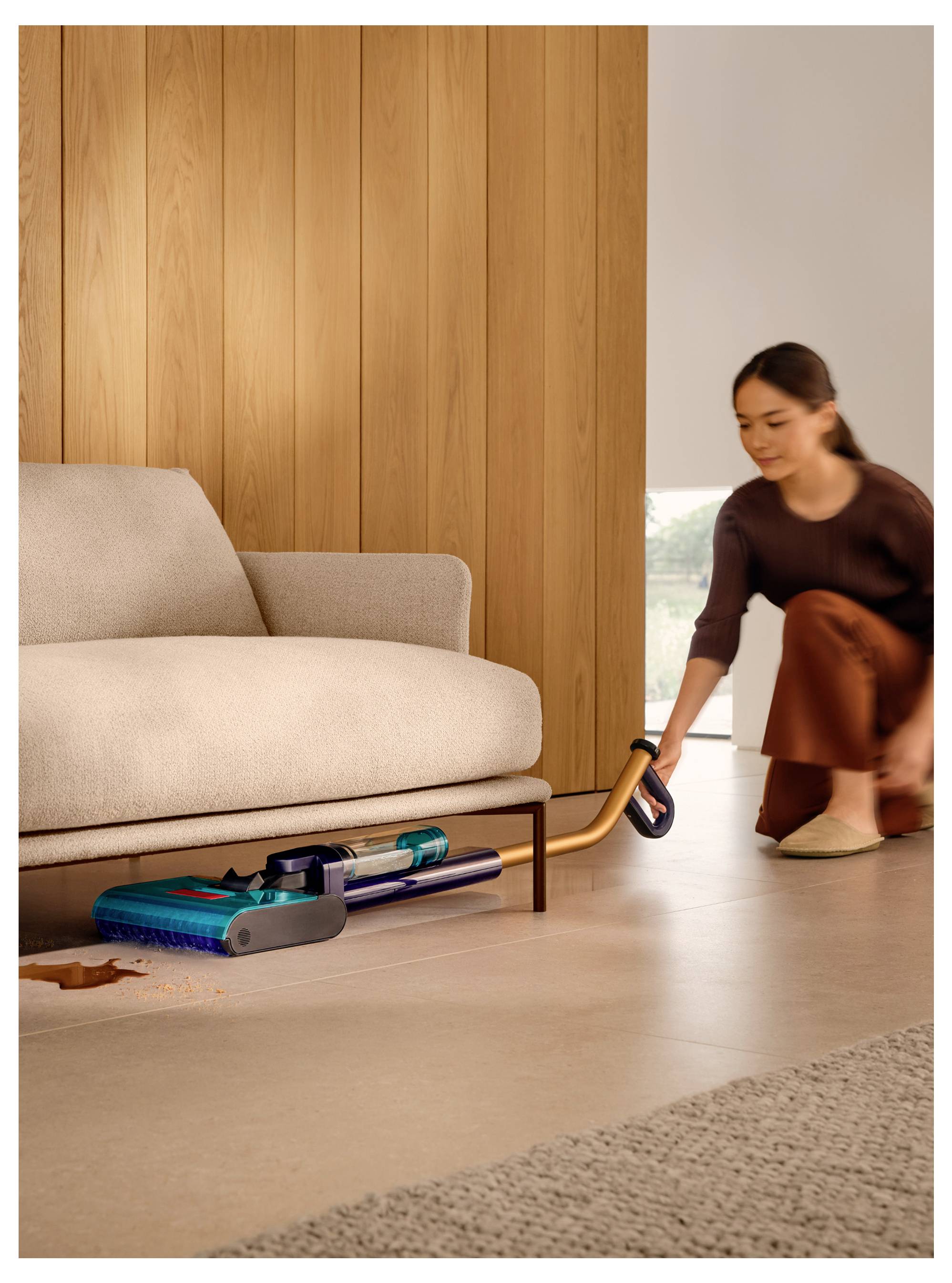 Eine Frau kniet neben einem Sofa und nutzt einen kabellosen Staubsauger, um Krümel vom Boden in einem modernen Wohnzimmer zu reinigen.