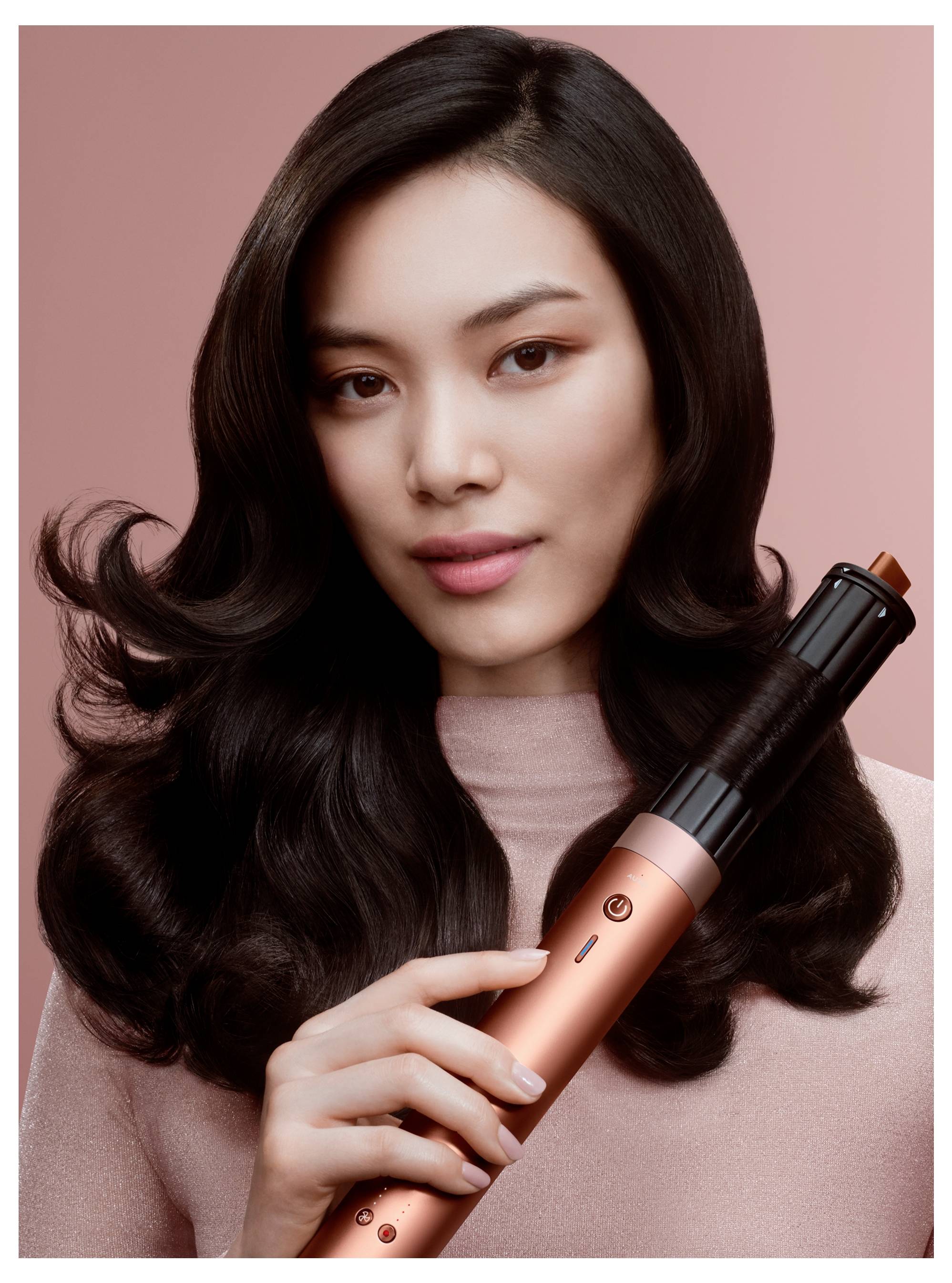 Frau mit gestyIten dunklen Haaren, die ein roségold- und schwarzes Lockenstab gegen einen rosa Hintergrund hält, was auf eine Schönheits- oder Haarstyling-Produktanzeige hindeutet.