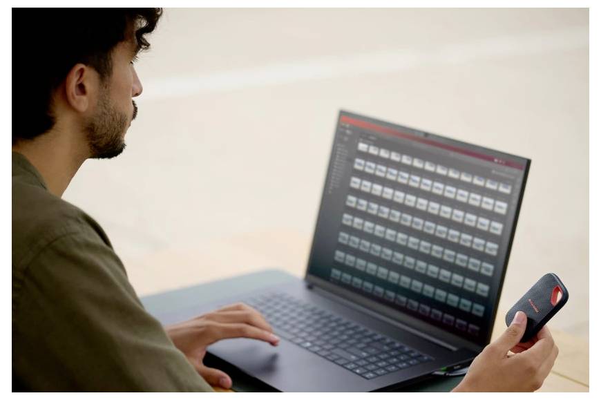 Mann mit einem Laptop, der ein Smartphone in der Hand hält und ein Raster von Dateien oder Miniaturansichten auf dem Laptop-Bildschirm überprüft.