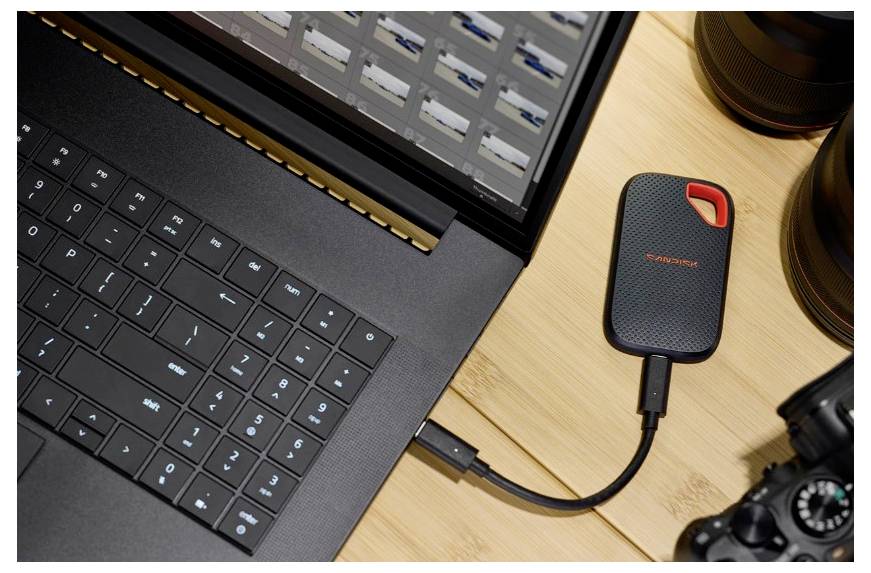 Laptop, der mit einer tragbaren externen SanDisk-SSD auf einem Schreibtisch verbunden ist, gezeigt neben Kameraausrüstung und einem Monitor.