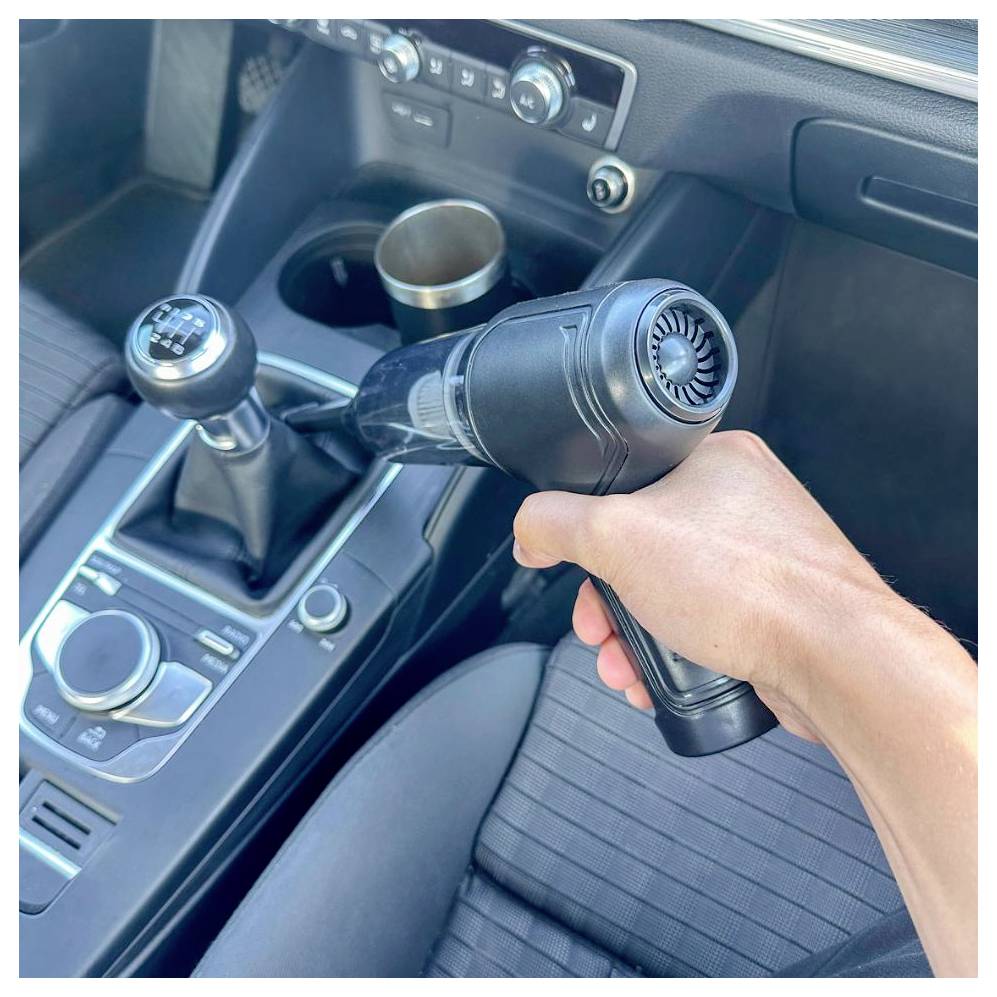 Eine Hand hält eine Heißluftpistole, die auf das Armaturenbrett eines Autos in der Nähe des Schaltgetriebes und des Getränkehalters gerichtet ist, was auf Innenreinigung oder Detaillierung hindeutet.