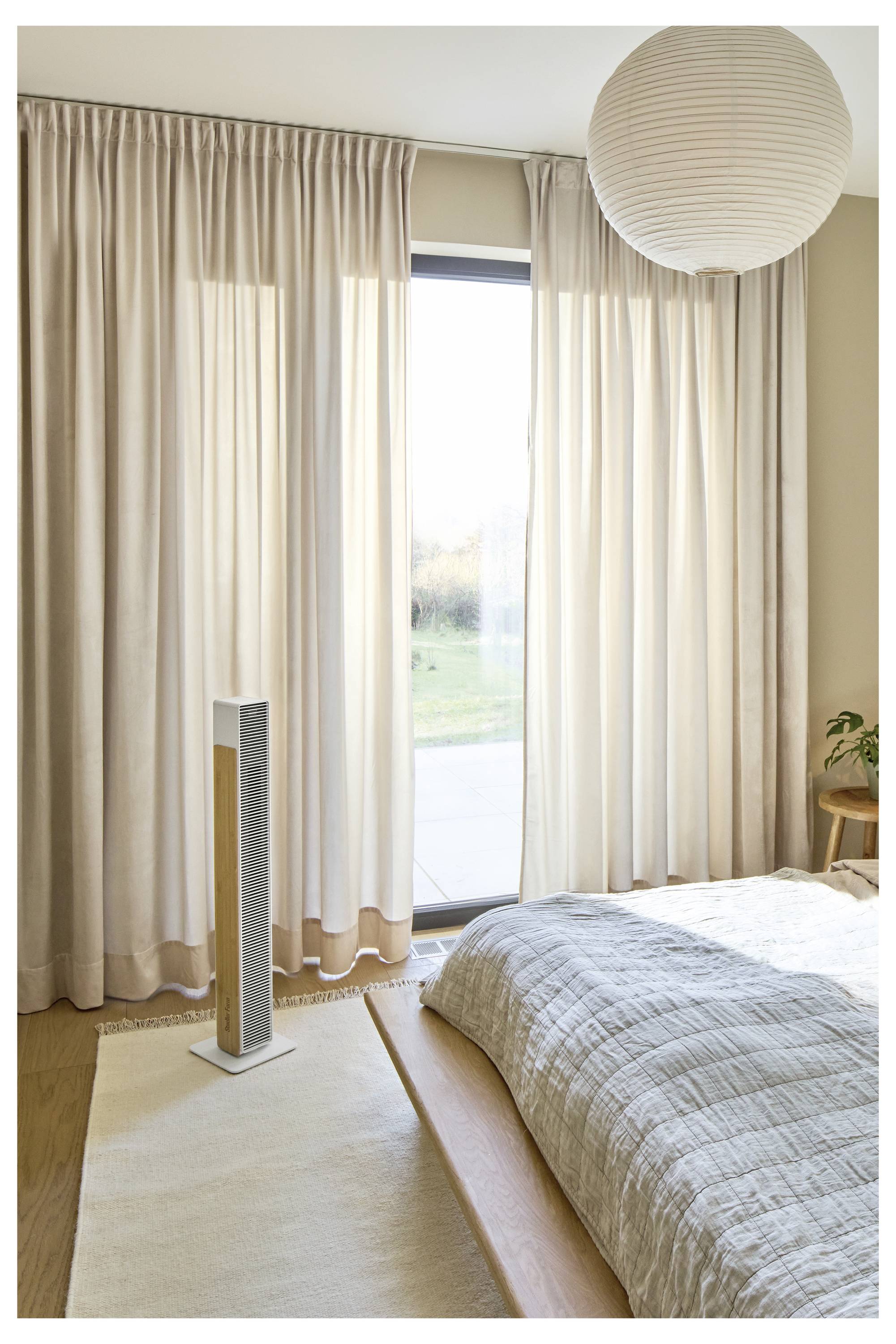 Helles Schlafzimmer mit beigen Vorhängen, die ein hohes Fenster bedecken, ein Bett mit grauer Steppdecke, ein Teppich, eine Stehlampe und eine Papierpendelleuchte.