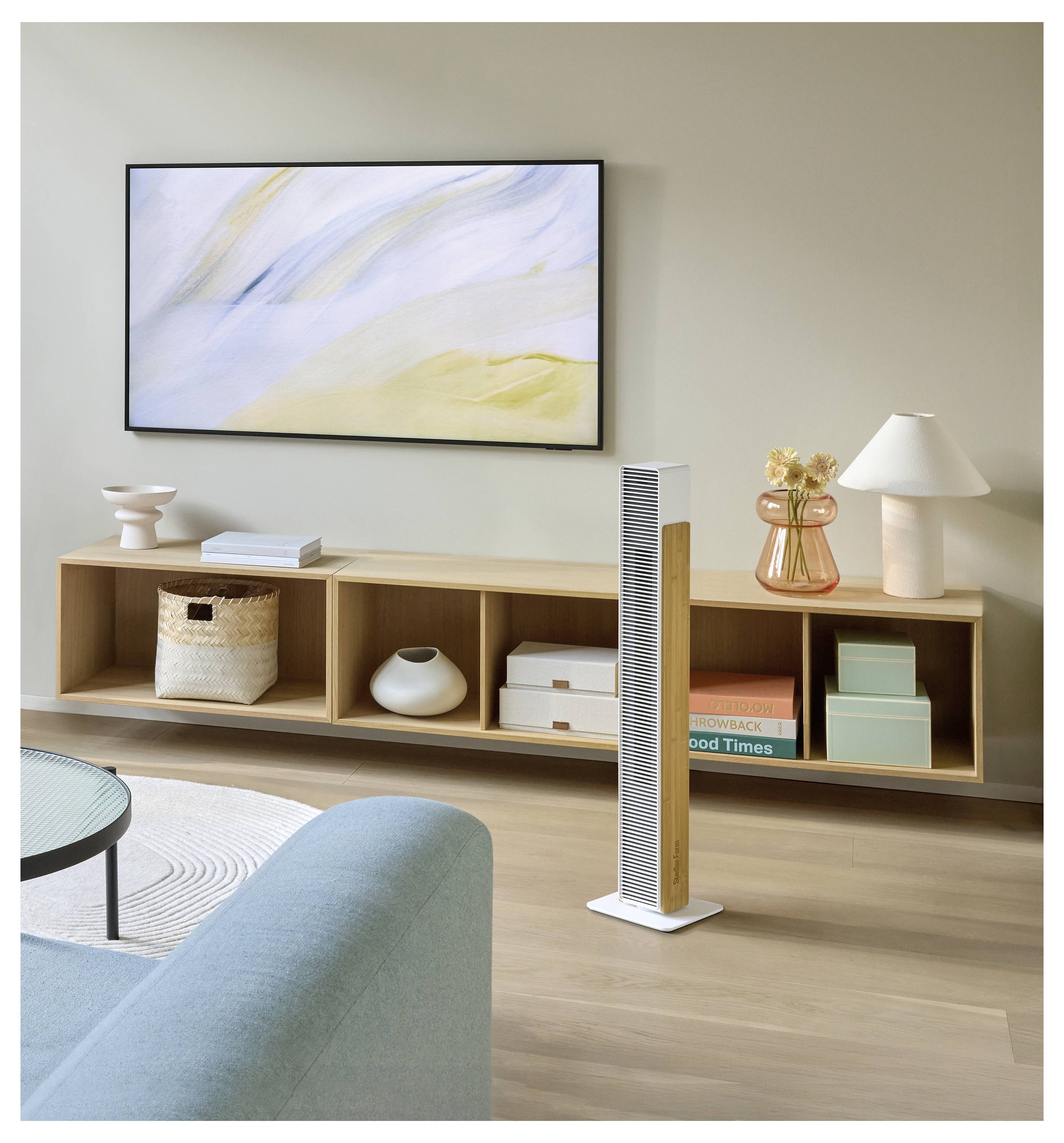 Modernes Wohnzimmer mit einem an der Wand montierten abstrakten Gemälde, niedrigem Holzregal, Lampe, Vase und einem hohen weißen Turm-Luftreiniger im Vordergrund.