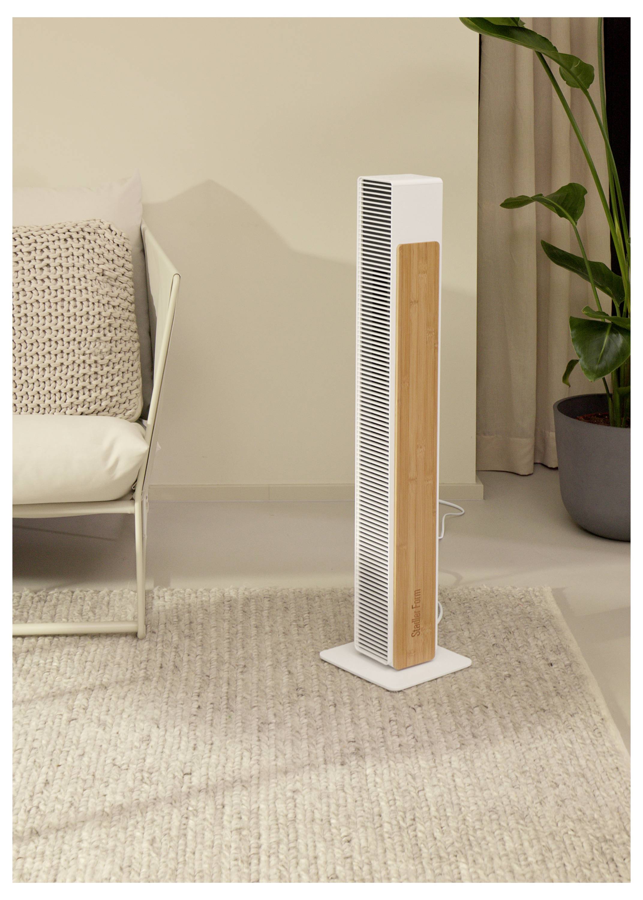 Großer weißer Turmventilator mit Holzakzent steht auf einem Teppich in einem Wohnzimmer neben einem Stuhl und einer Topfpflanze.