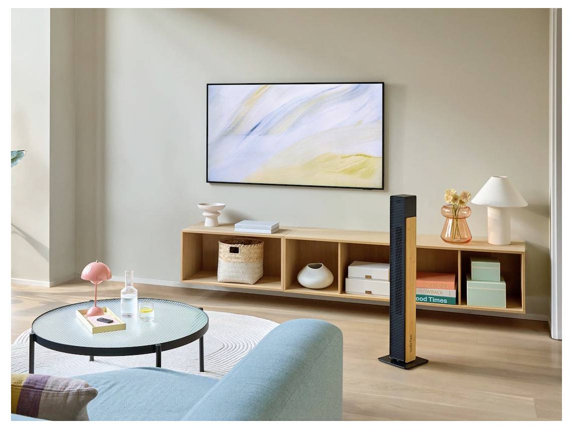 Ein gemütliches modernes Wohnzimmer mit einem Sofa, einem runden Couchtisch, einer niedrigen Holzkonsole mit Dekor, Wandkunst und einem schlanken Turmluftreini­ger neben den Regalen.