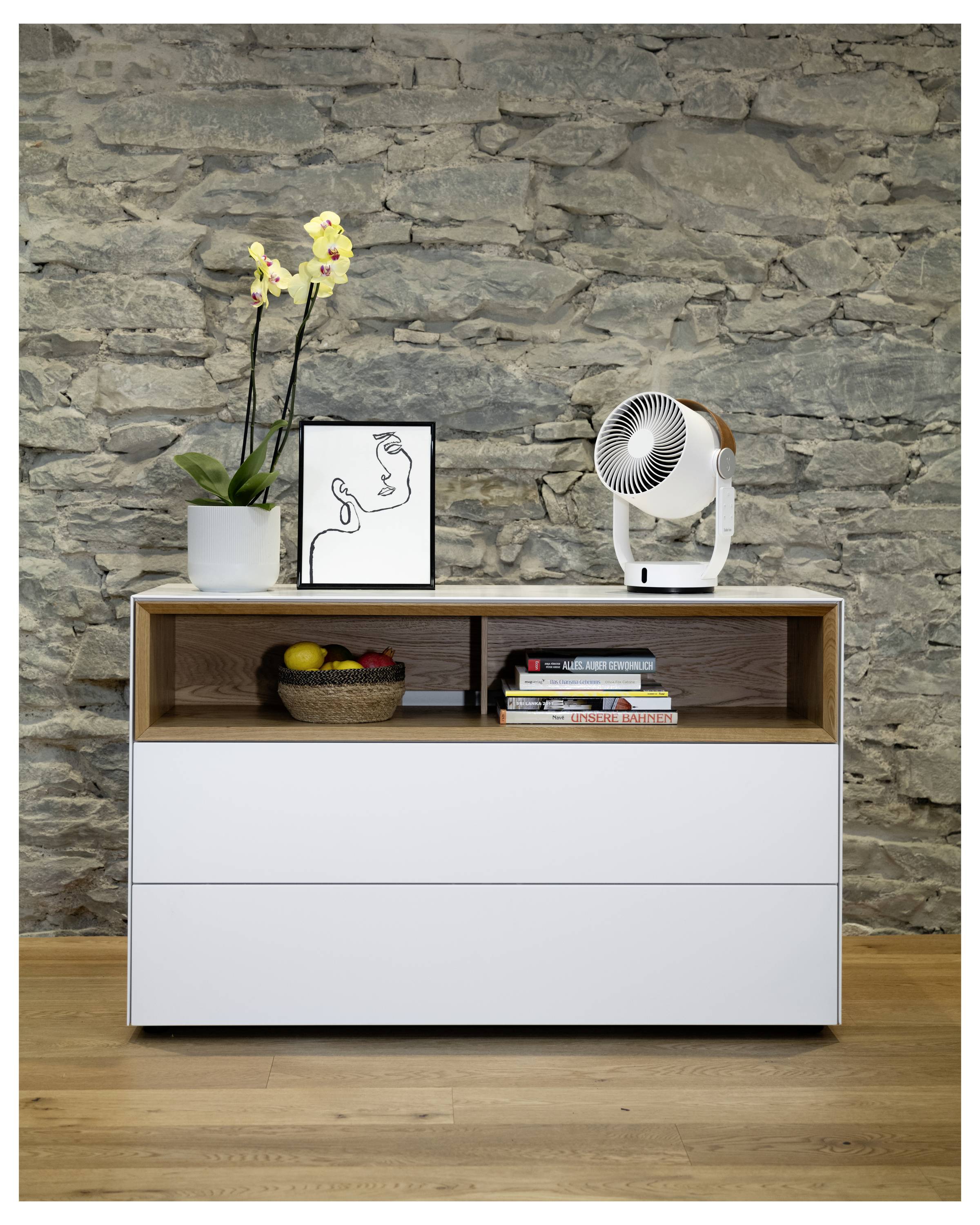 Moderner Kommode an einer Steinmauer, oben mit einer Topforchidee, gerahmter Linienkunst und einem kleinen Ventilator; offenes Regal mit Büchern und einer Obstschale.