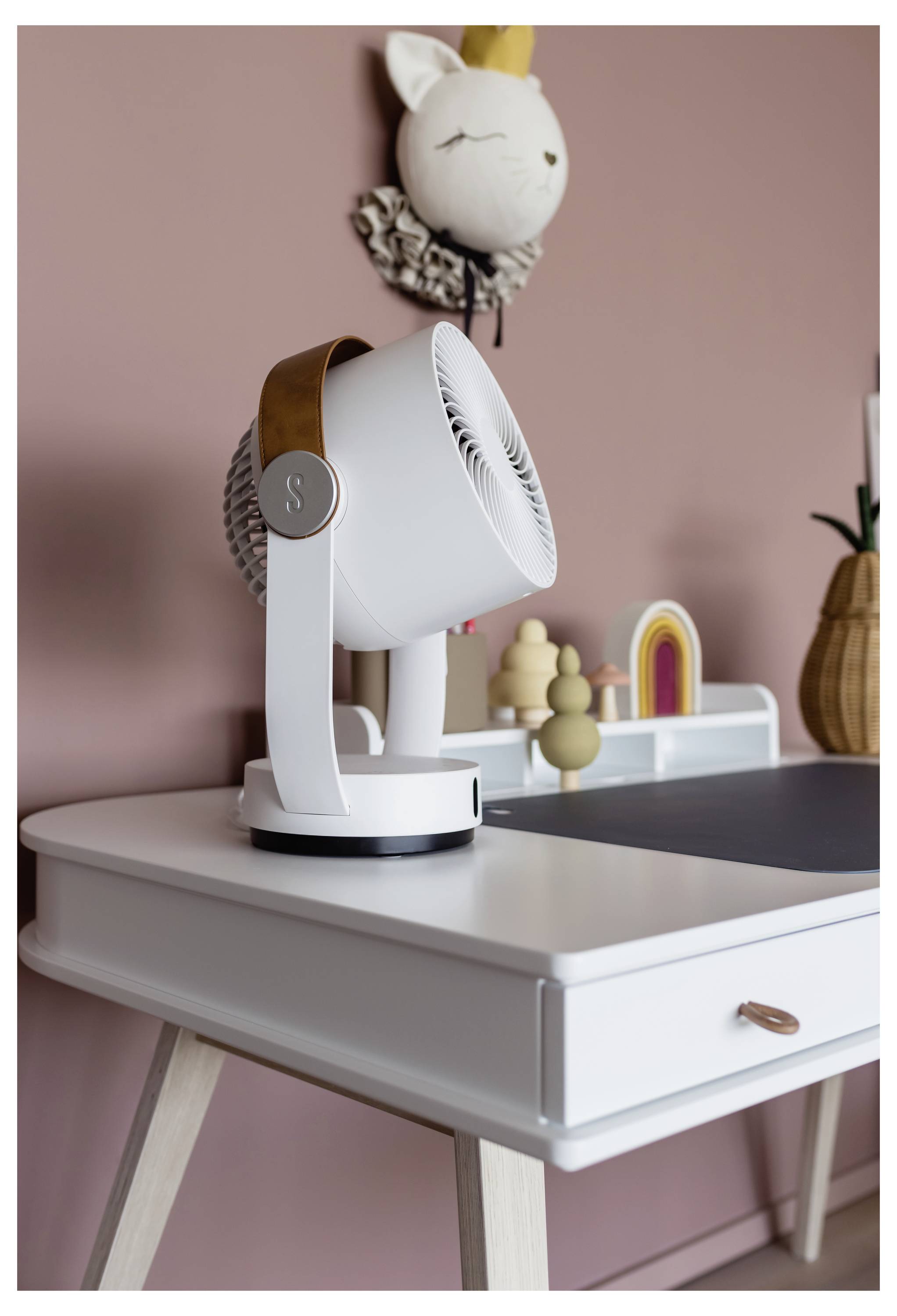Weißer Schreibtischventilator auf einem Schreibtisch aus hellem Holz in einem rosa Zimmer, mit einem kleinen Regal und weicher Wanddekoration dahinter.