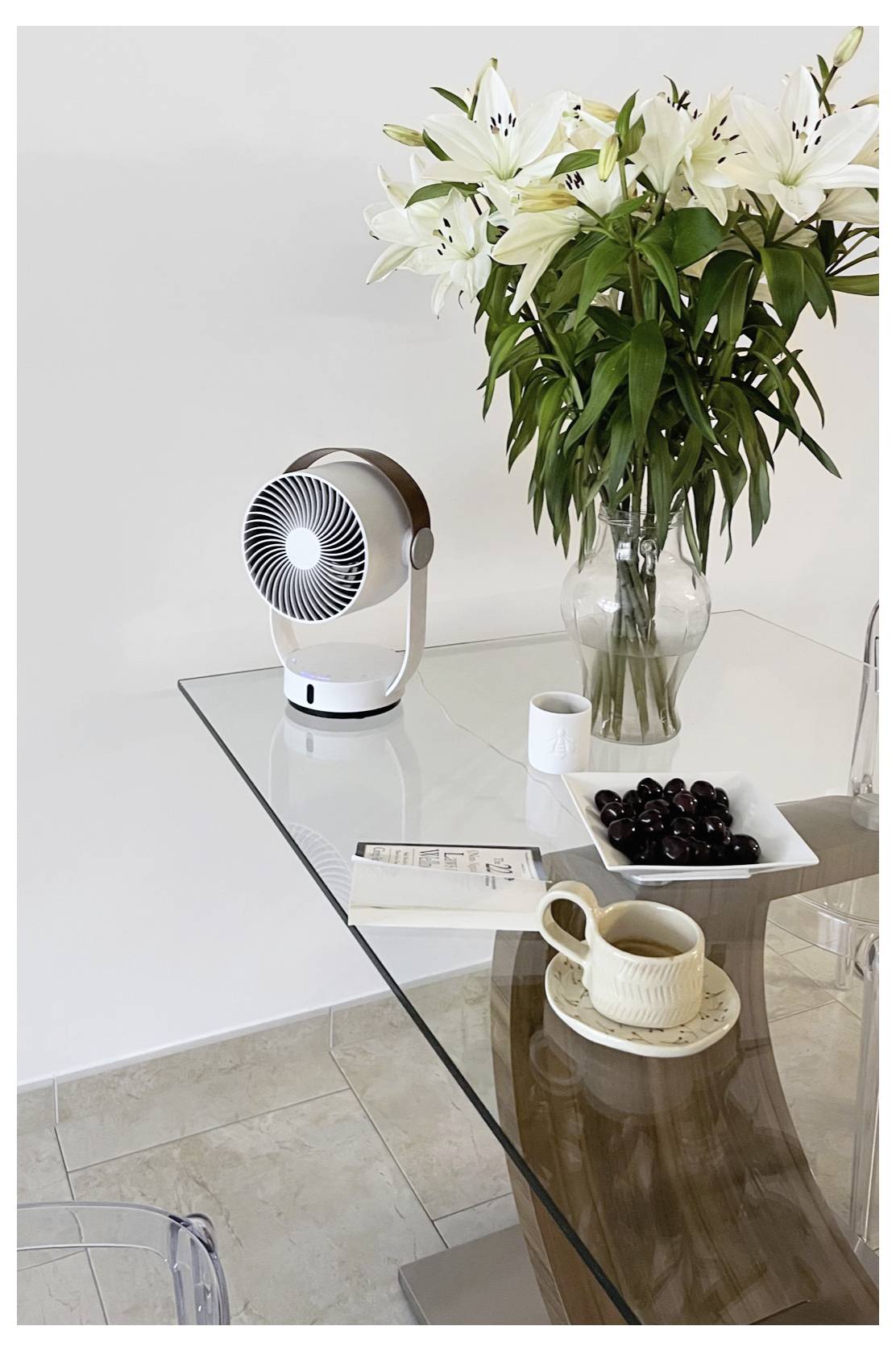 Glasesstisch mit weißem Ventilator, Vase mit weißen Lilien, Kaffeetasse und Obstschale in einem hellen, minimalistischen Rauminterieur.