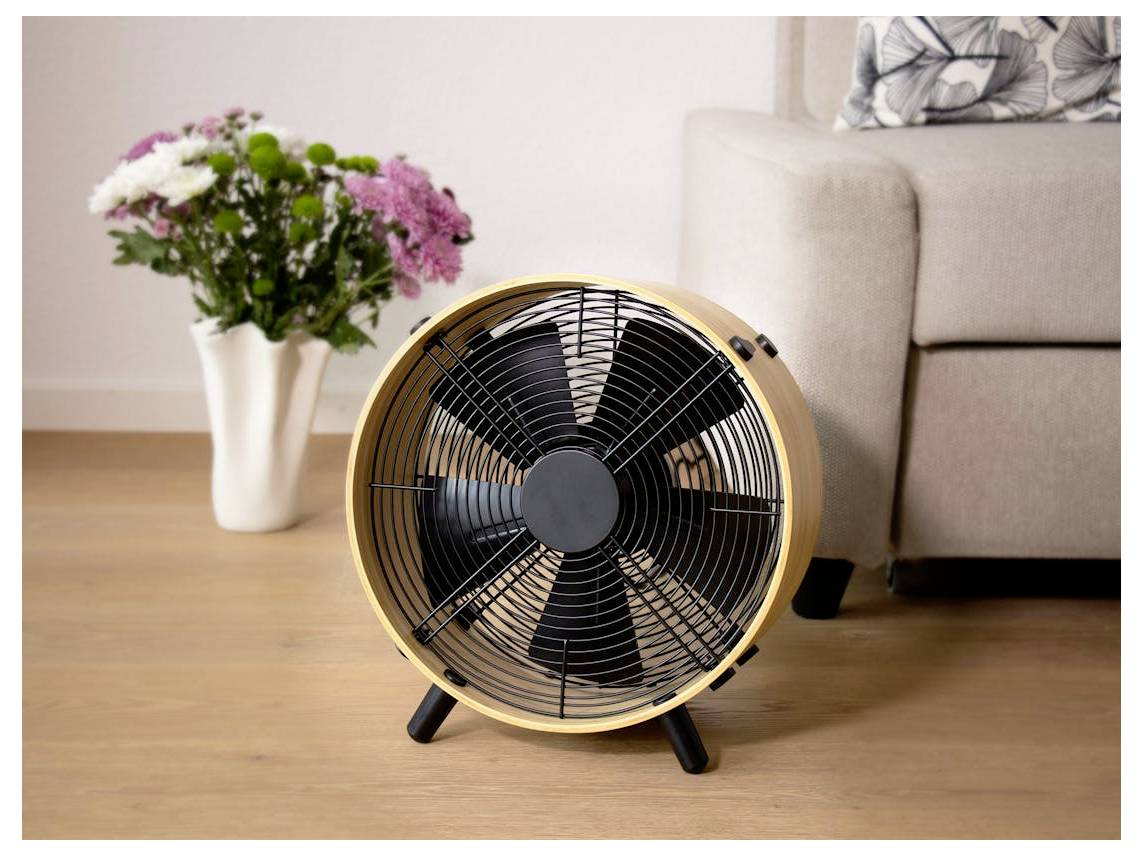 Ein stilvolles Wohnzimmer mit einem Holzventilator auf dem Boden, einem beigefarbenen Sofa mit Kissen, einem Couchtisch, einer großen Pflanze und einem gewebten Pouf am Fenster.