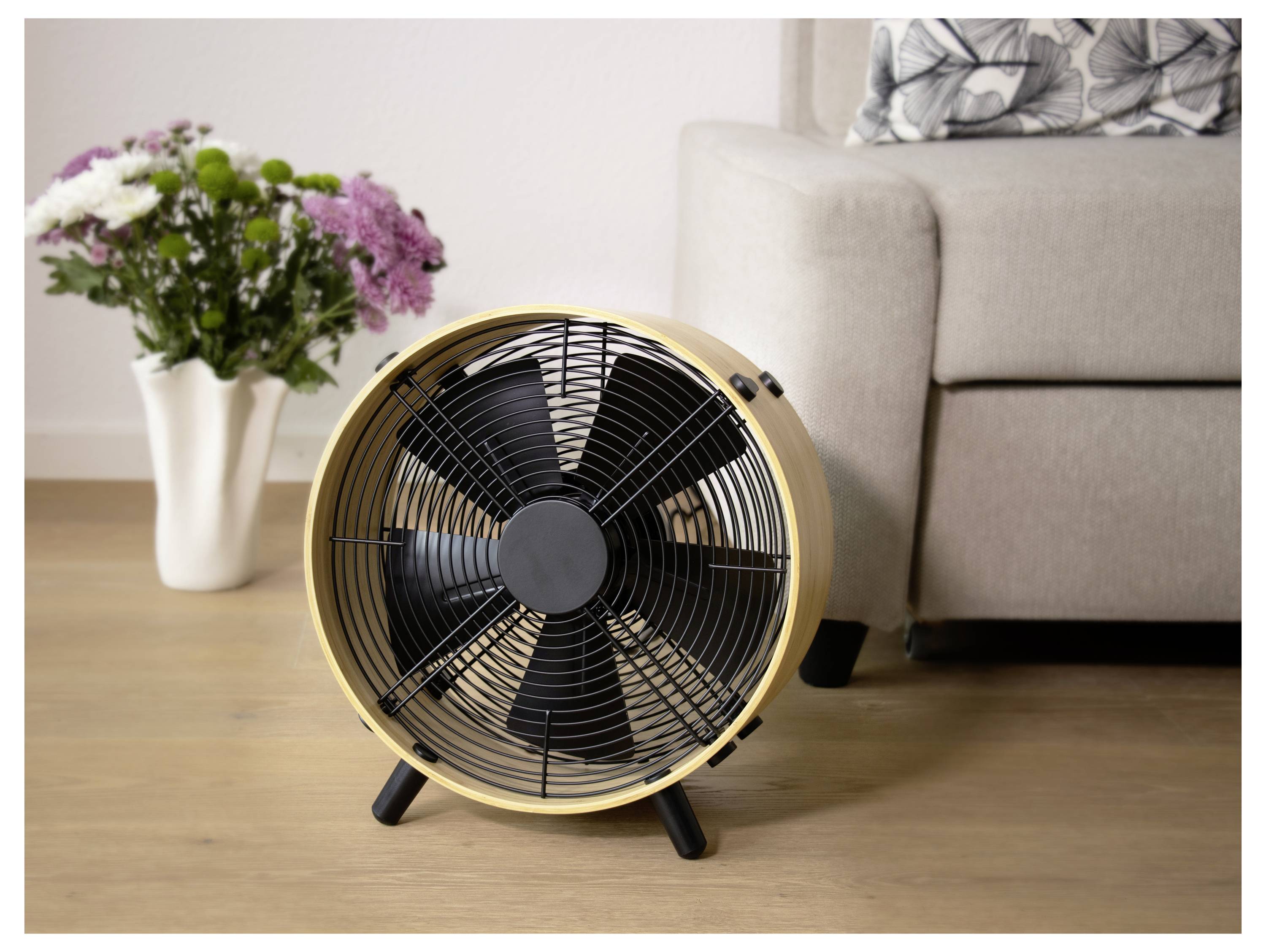 Kleiner Tischventilator auf einem Holzboden in einem Wohnzimmer neben einem Sofa und einer Vase mit Blumen.