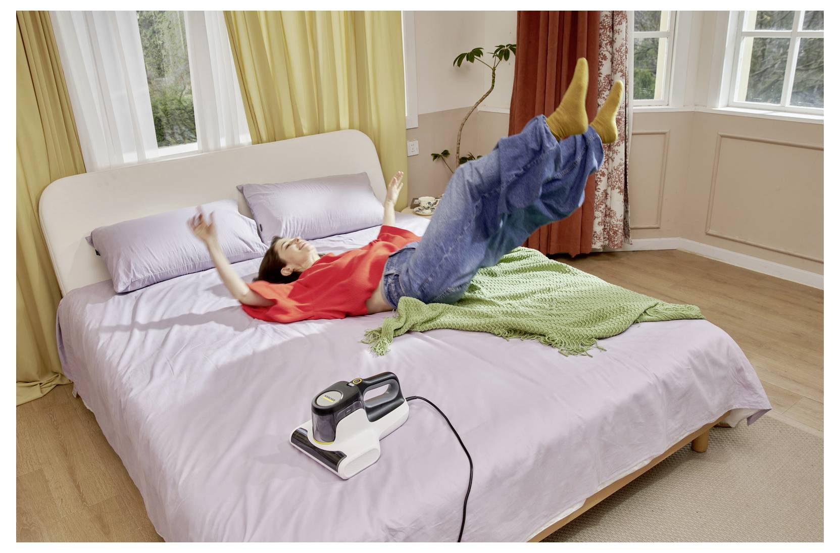 Eine Person liegt auf einem Bett und benutzt eine vibrierende Trainingsmaschine, während ihre Beine angehoben sind und eine grüne Decke neben ihr in einem hellen Schlafzimmer liegt.