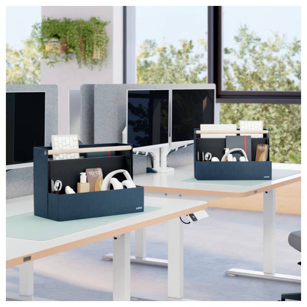 Moderne Bürotische mit schwarzen Caddy-Organizern, die Kopfhörer, Flaschen, Notizbücher und Tastaturen neben Computermonitoren bei großen Fenstern halten.