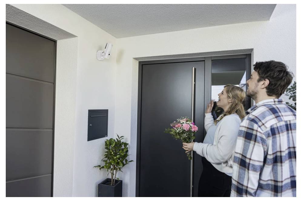 Ein Paar steht vor einer modernen Apartmenttür, die Frau hält rosa Blumen und läutet die Türklingel.