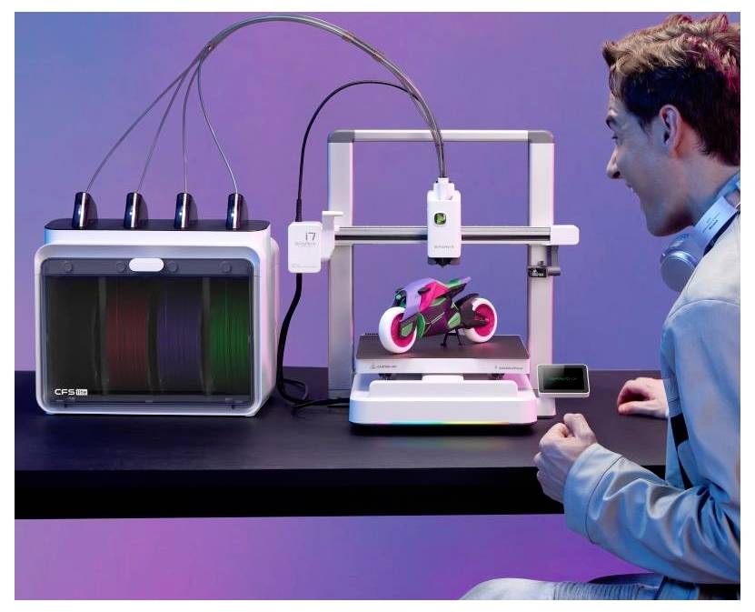 Ein Mann beobachtet, wie ein 3D-Drucker ein kleines buntes Motorradmodell neben einer verbundenen Druckereinheit auf einem Tisch anzeigt.