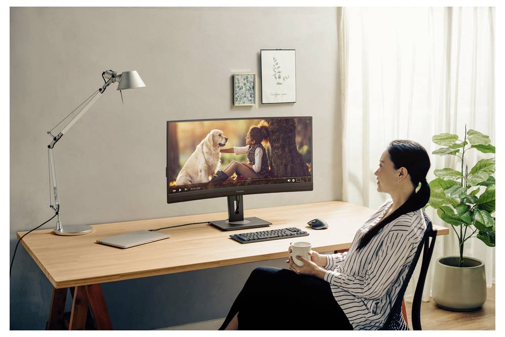 Frau sitzt an einem Schreibtisch zu Hause, hält eine Tasse in der Hand und schaut auf einen Monitor, auf dem eine Frau mit einem Hund zu sehen ist; Laptop, Tastatur, Lampe und eine Pflanze befinden sich auf dem Schreibtisch.