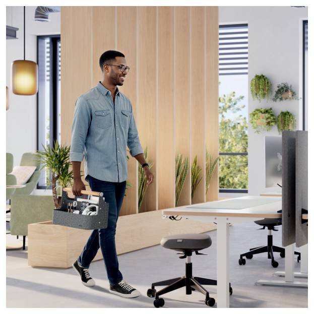 Ein Mann trägt eine Werkzeugkiste durch ein modernes Büro in Richtung Schreibtische und Monitore und deutet dabei auf Einrichtungs- oder Wartungsarbeiten hin.