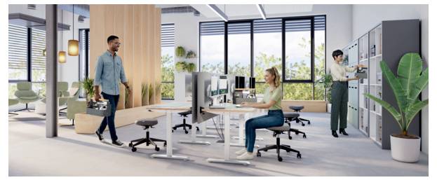 Großraumbüro mit einem Mann, der eine Aktentasche trägt und an einer sitzenden Frau vorbeigehen, die an einem Computer arbeitet, mit Schreibtischen, Pflanzen und Spinden im Hintergrund.