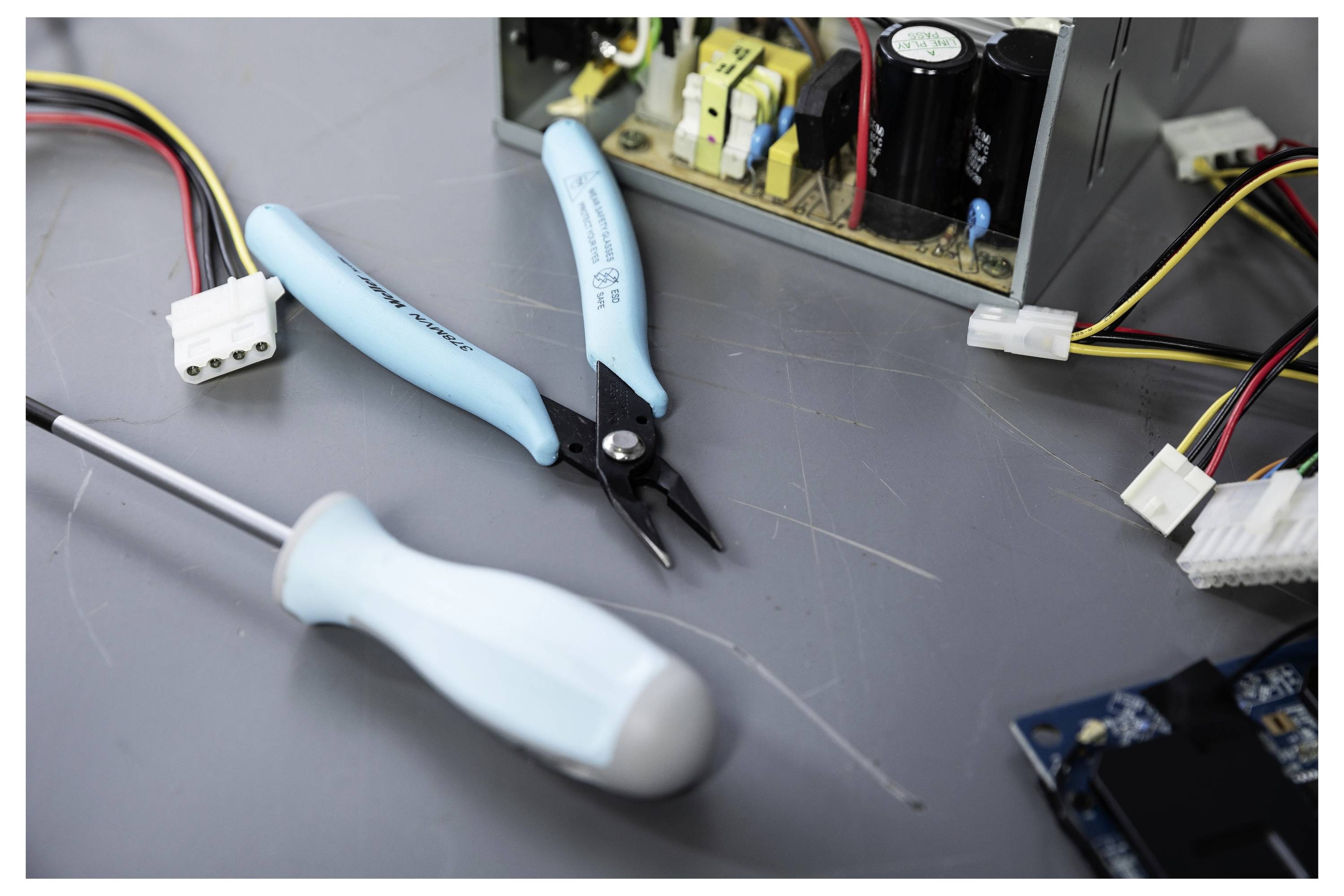 Nahaufnahme von blaugriffigen elektronischen Reparaturwerkzeugen, einschließlich Zangen und eines Schraubenziehers, auf einer Werkbank neben Computerhardware und Kabeln.