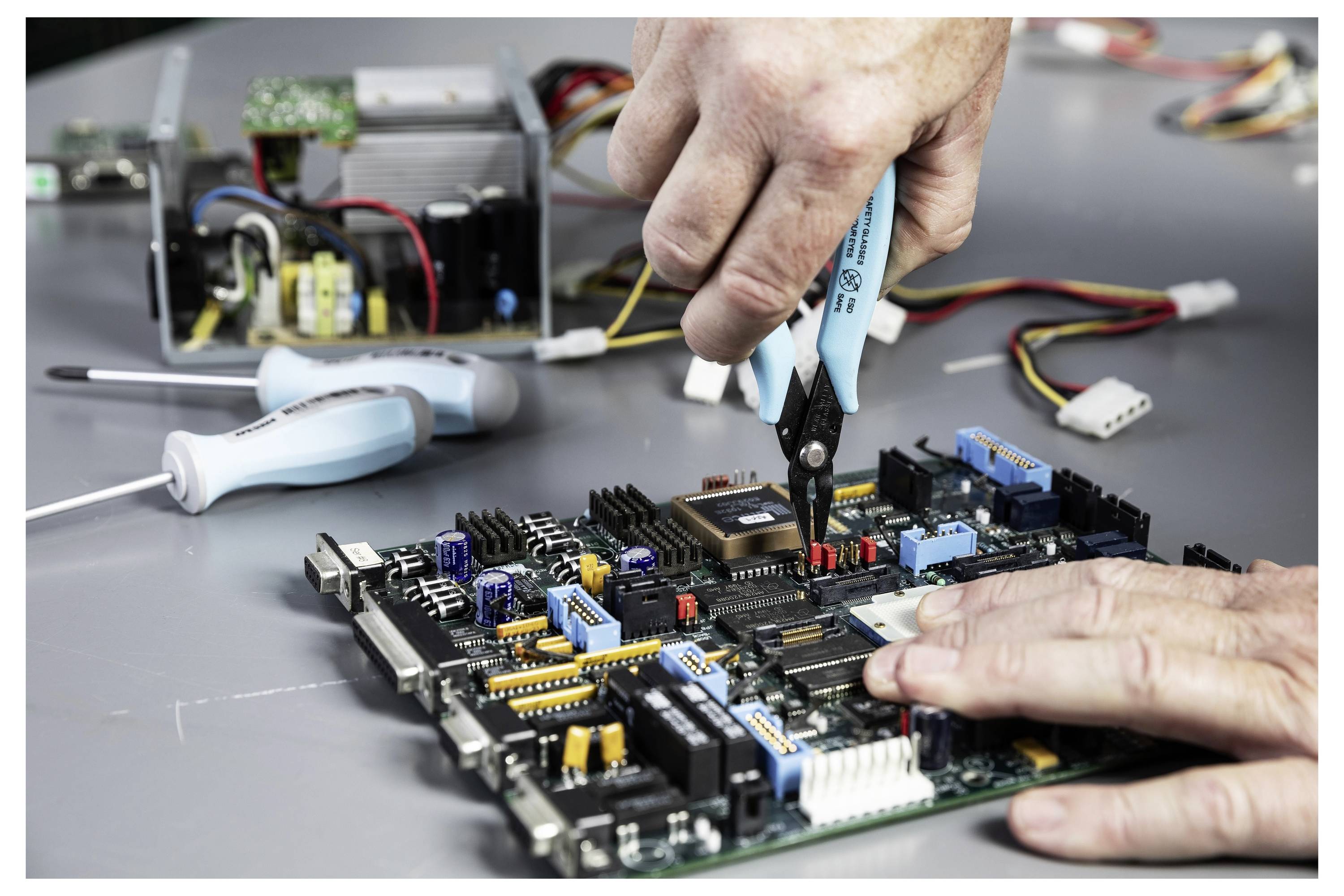 Ein Techniker nutzt eine Zange, um Komponenten auf einer Leiterplatte zu reparieren oder zu verbinden, auf einer Arbeitsbank mit Testwerkzeugen und Drähten.