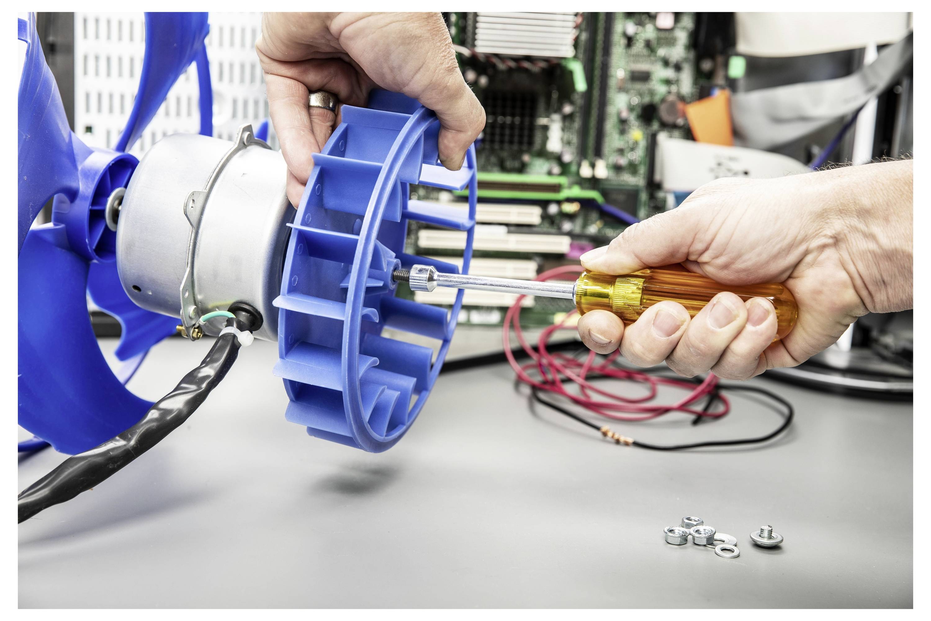 Hände, die einen Schraubendreher verwenden, um eine blaue Elektromotor- oder Lüfterkomponente auf einer Werkbank zusammenzusetzen oder zu reparieren, mit Werkzeugen und Drähten in der Nähe.