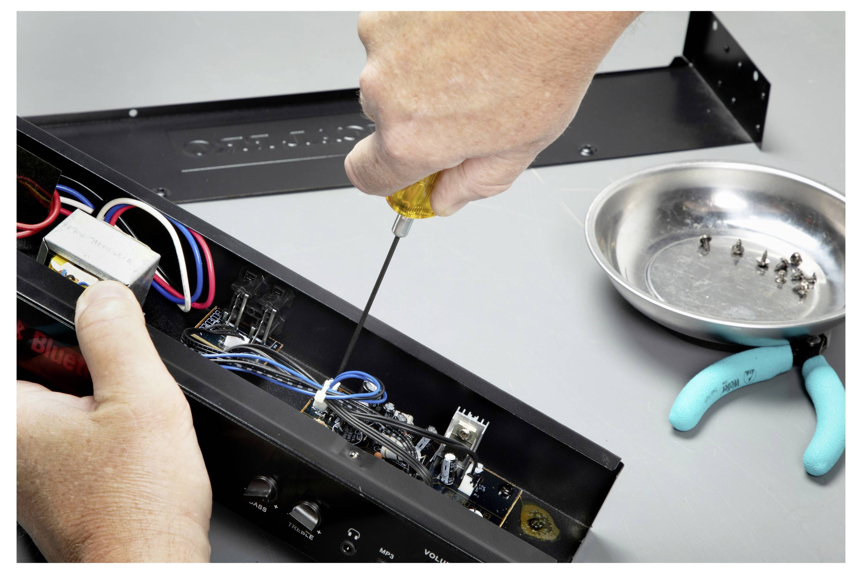 Hände verwenden einen Schraubendreher zur Reparatur des Inneren eines elektronischen Geräts, mit freiliegenden Drähten und kleinen Teilen in einer nahegelegenen Schale.