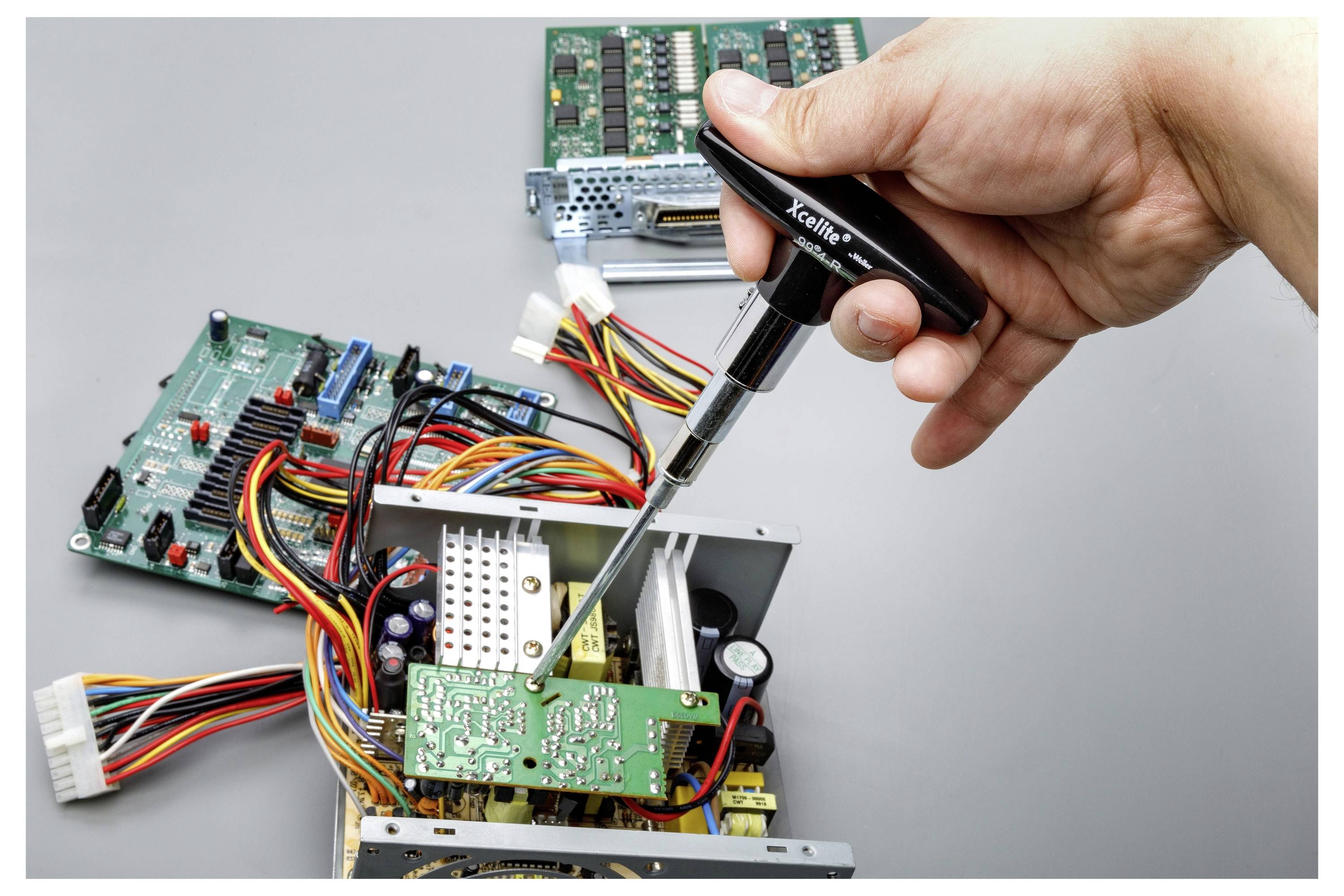 Nahaufnahme einer Hand, die einen Schraubenzieher verwendet, um eine elektronische Leiterplatte in einem geöffneten Gerät zu reparieren, mit freiliegenden Drähten und Komponenten.