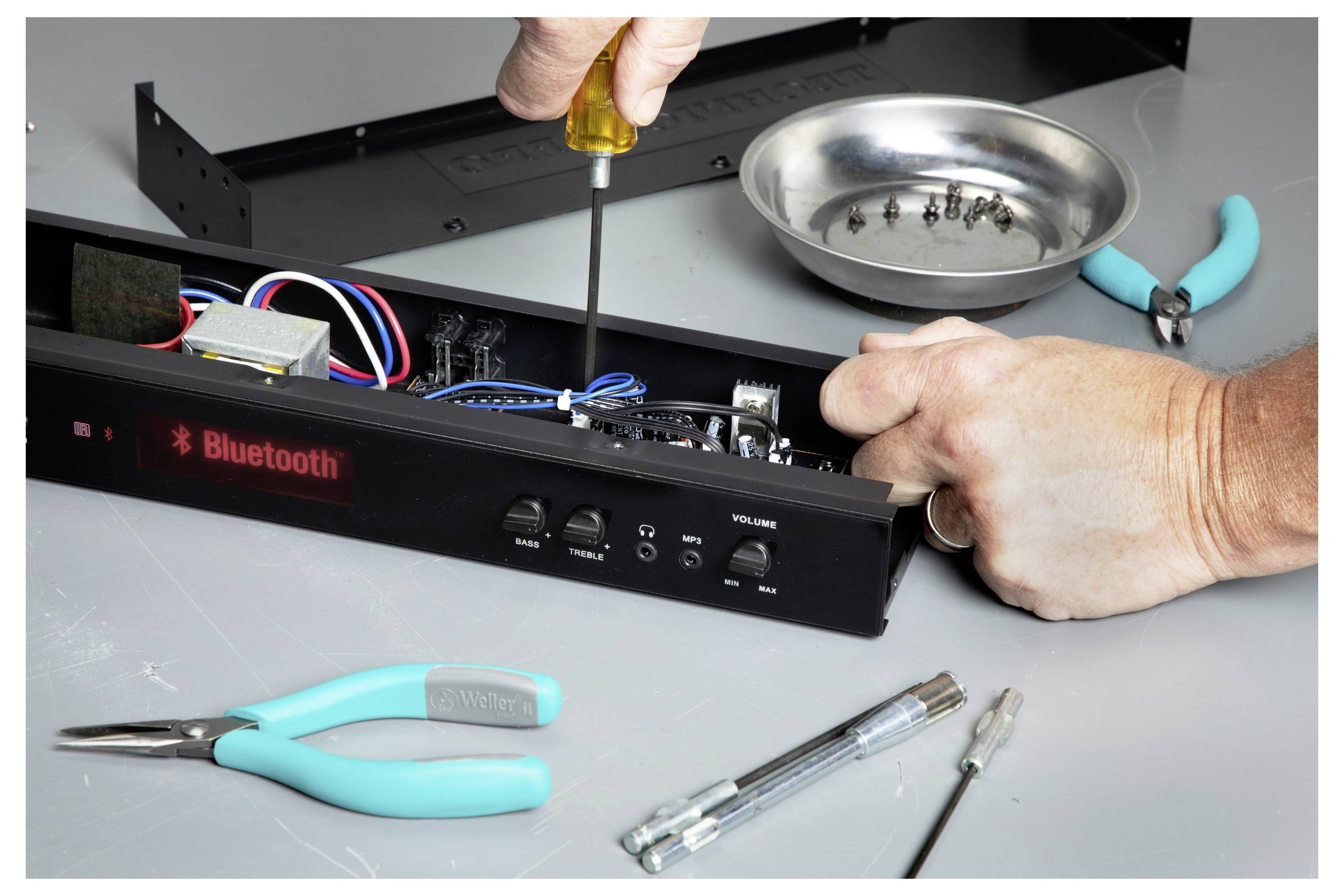 Techniker repariert ein Bluetooth-Audiogerät mit einem Schraubenzieher, umgeben von Werkzeugen und offenen Komponenten auf einer Werkbank.