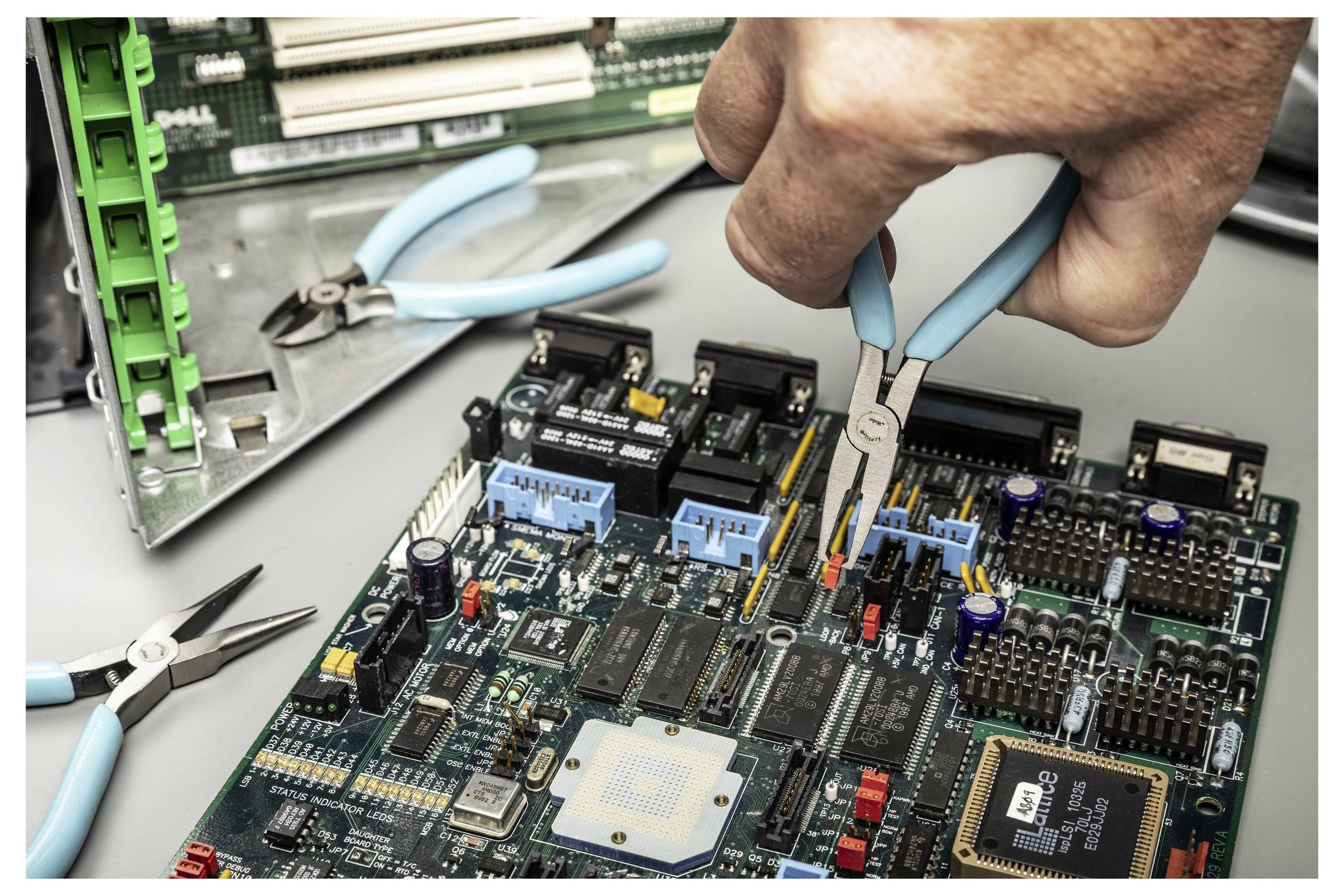 Hand mit Spitzenzange zur Anpassung von Teilen auf einer Leiterplatte während der Elektronikreparatur, mit Werkzeugen und Computerhardware in der Nähe.