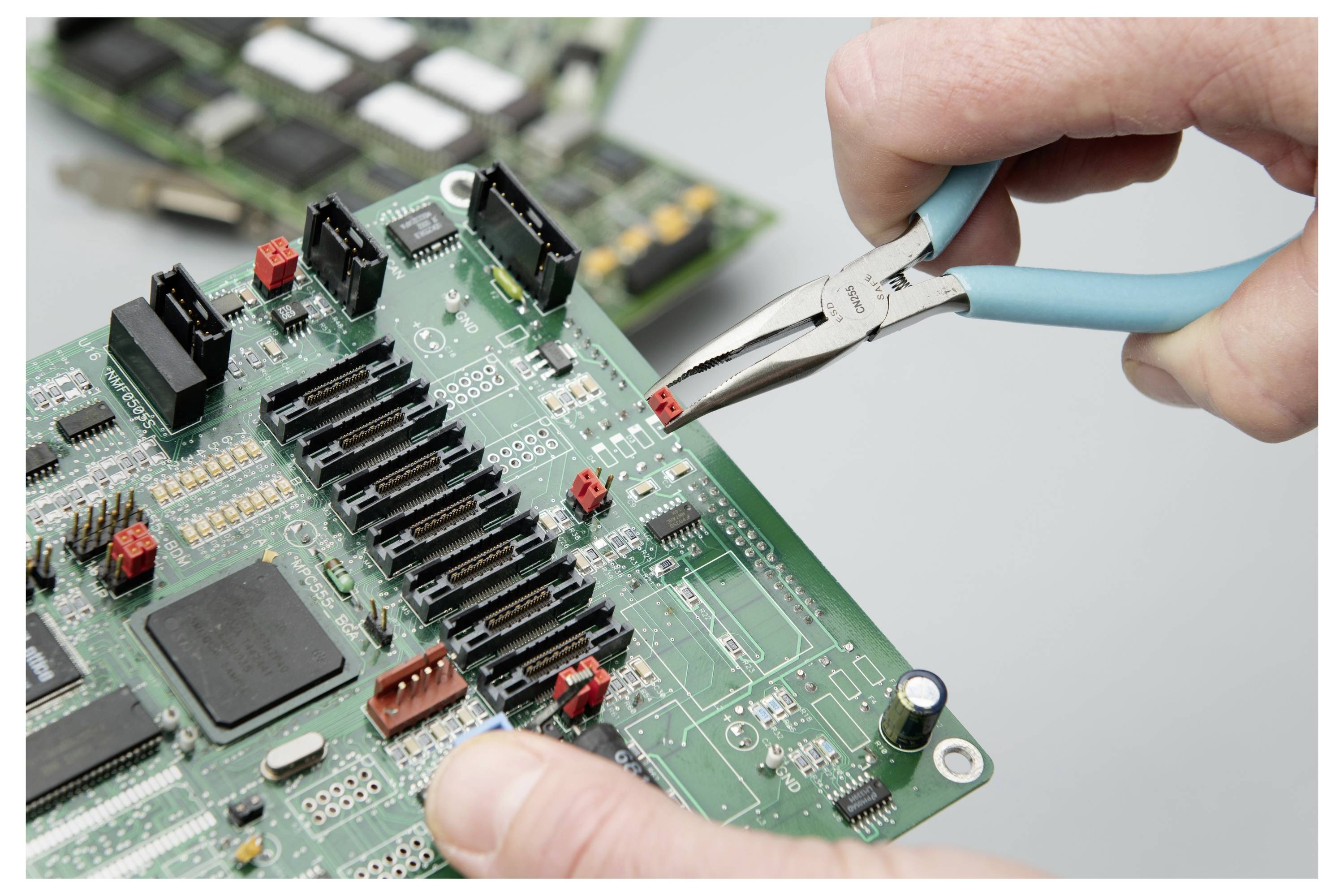 Hände halten eine Spitzenzange und entfernen ein kleines rotes Bauteil von einer grünen Leiterplatte während einer Elektronikreparatur.