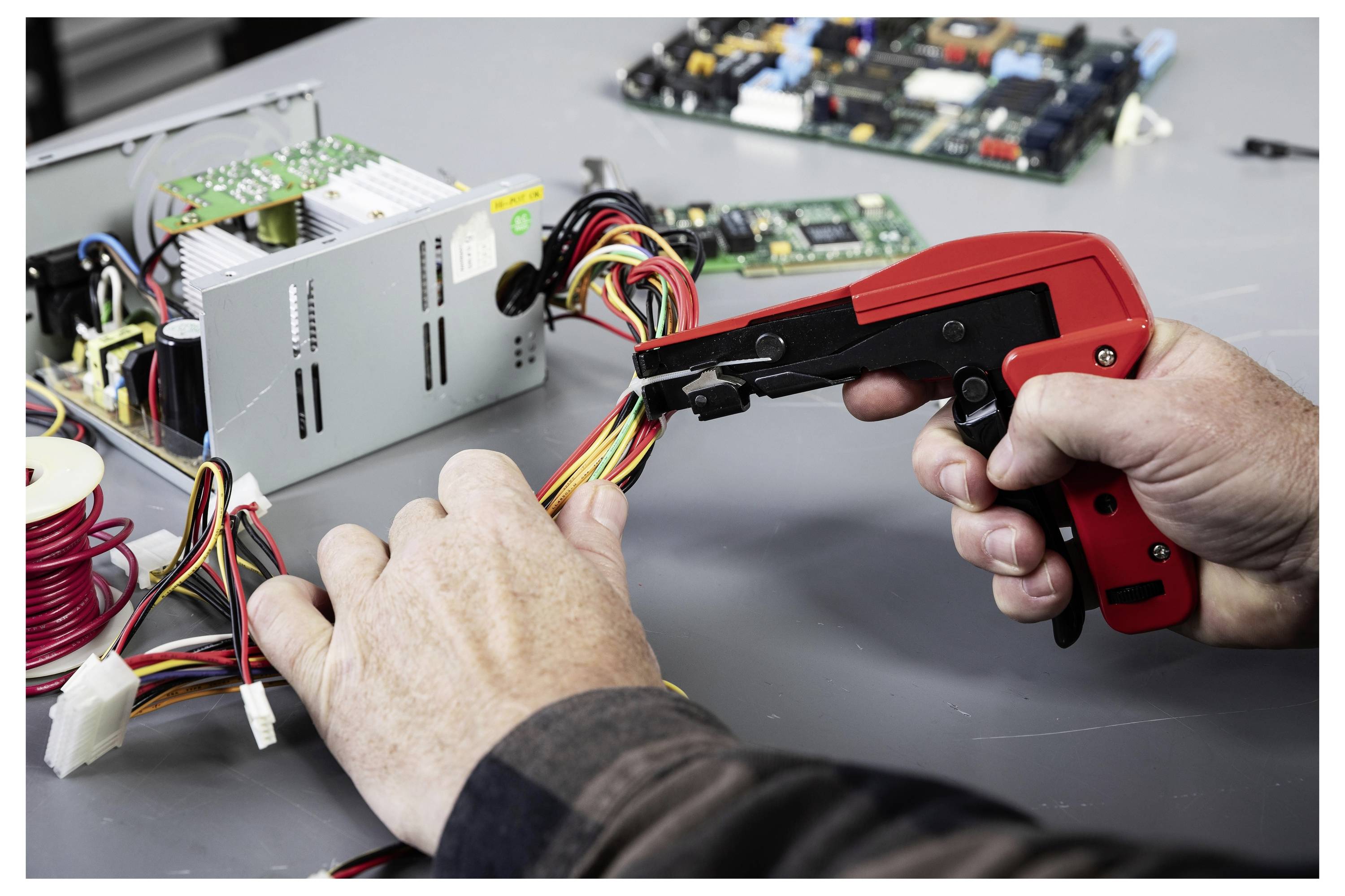 Hände, die einen roten Abisolierer verwenden, um mehrfarbige Computerkabel neben einem geöffneten Netzteil auf einer Werkbank zu schneiden und abzuisolieren.