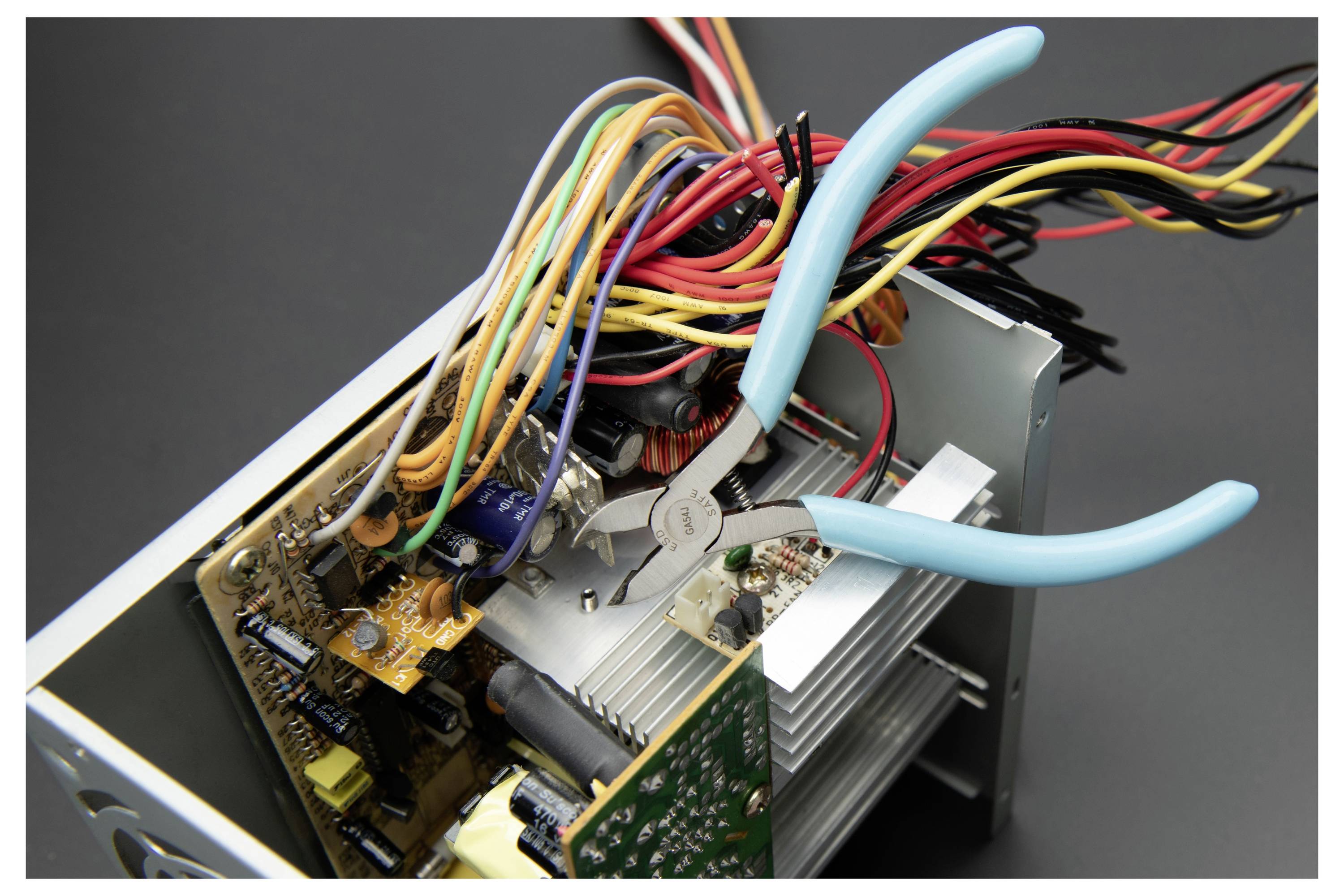 Geöffnetes elektronisches Gerät mit verwirrtem mehrfarbigem Drahtgeflecht und blauem Zanghengriff, der innen in einem Metallgehäuse arbeitet, was auf Reparatur oder Überprüfung hindeutet.
