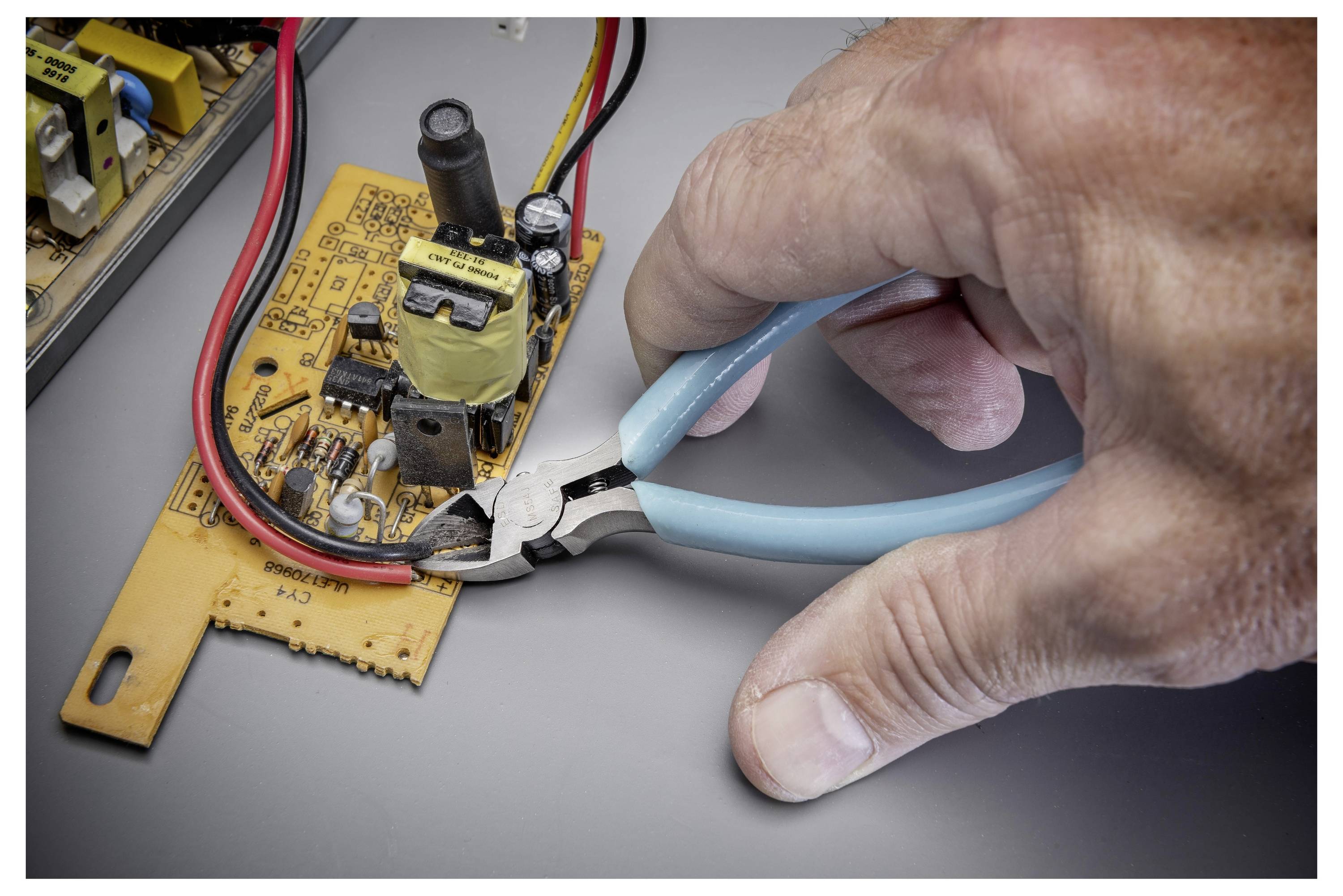 Hand mit blauen Schneidern, die einen Draht auf einer kleinen gelben Leiterplatte während einer Elektronikreparatur abschneidet.