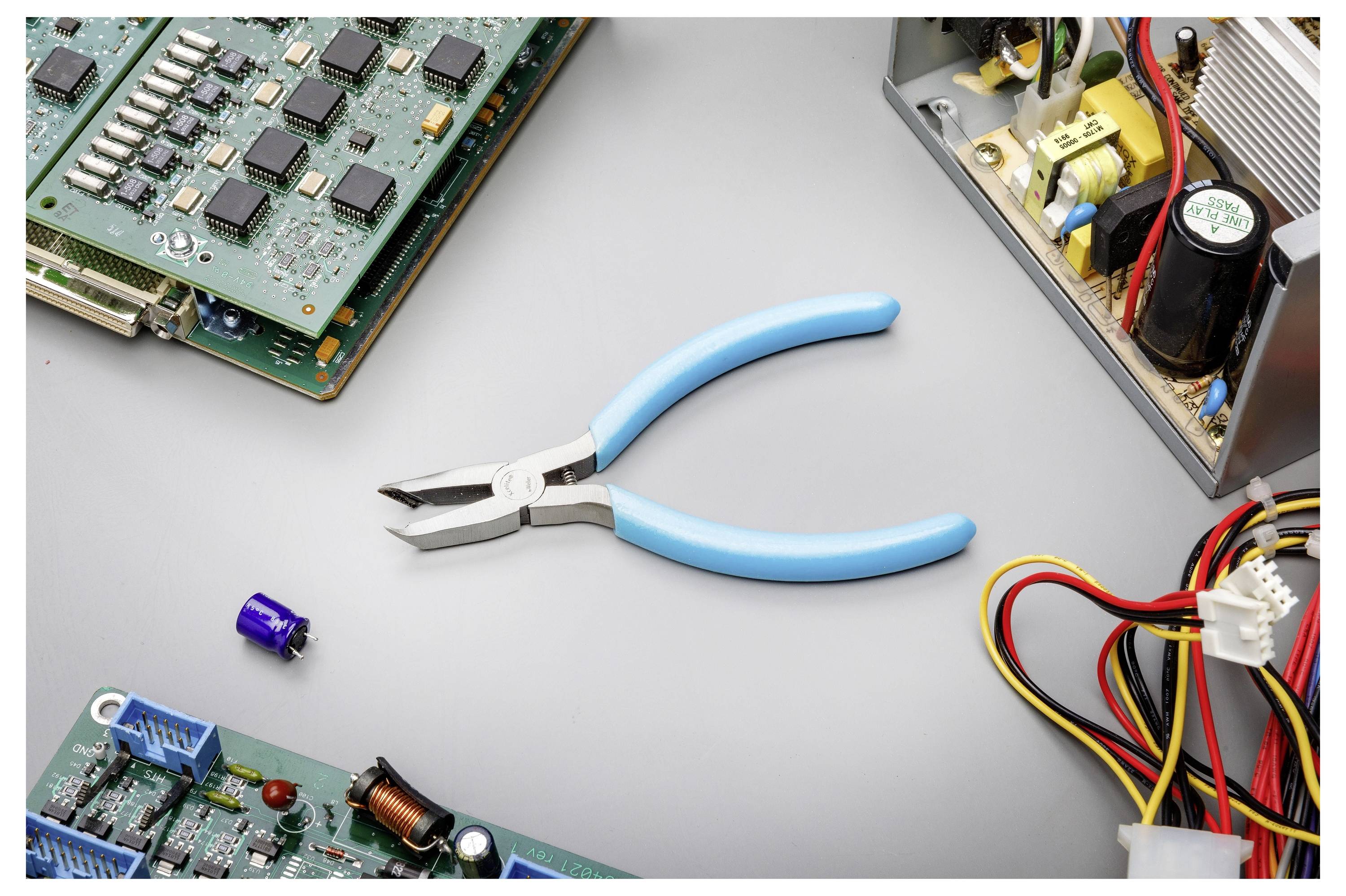 Blaugriffige Zange, die auf einer Werkbank zentriert ist, umgeben von Leiterplatten, Drähten und einem offenen elektronischen Gerät, was auf Elektronikreparatur oder -montage hindeutet.