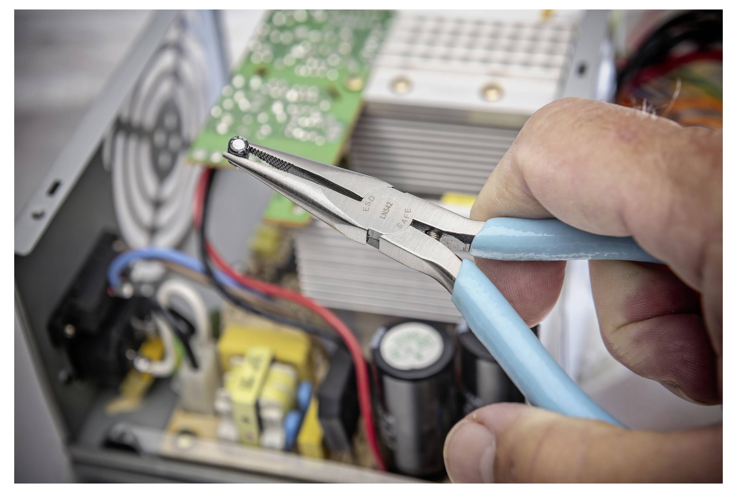 Hand hält eine Spitzenzange in einem offenen elektronischen Gerät und arbeitet in der Nähe von Leiterplatten, Verdrahtung und einem Kühlventilator.