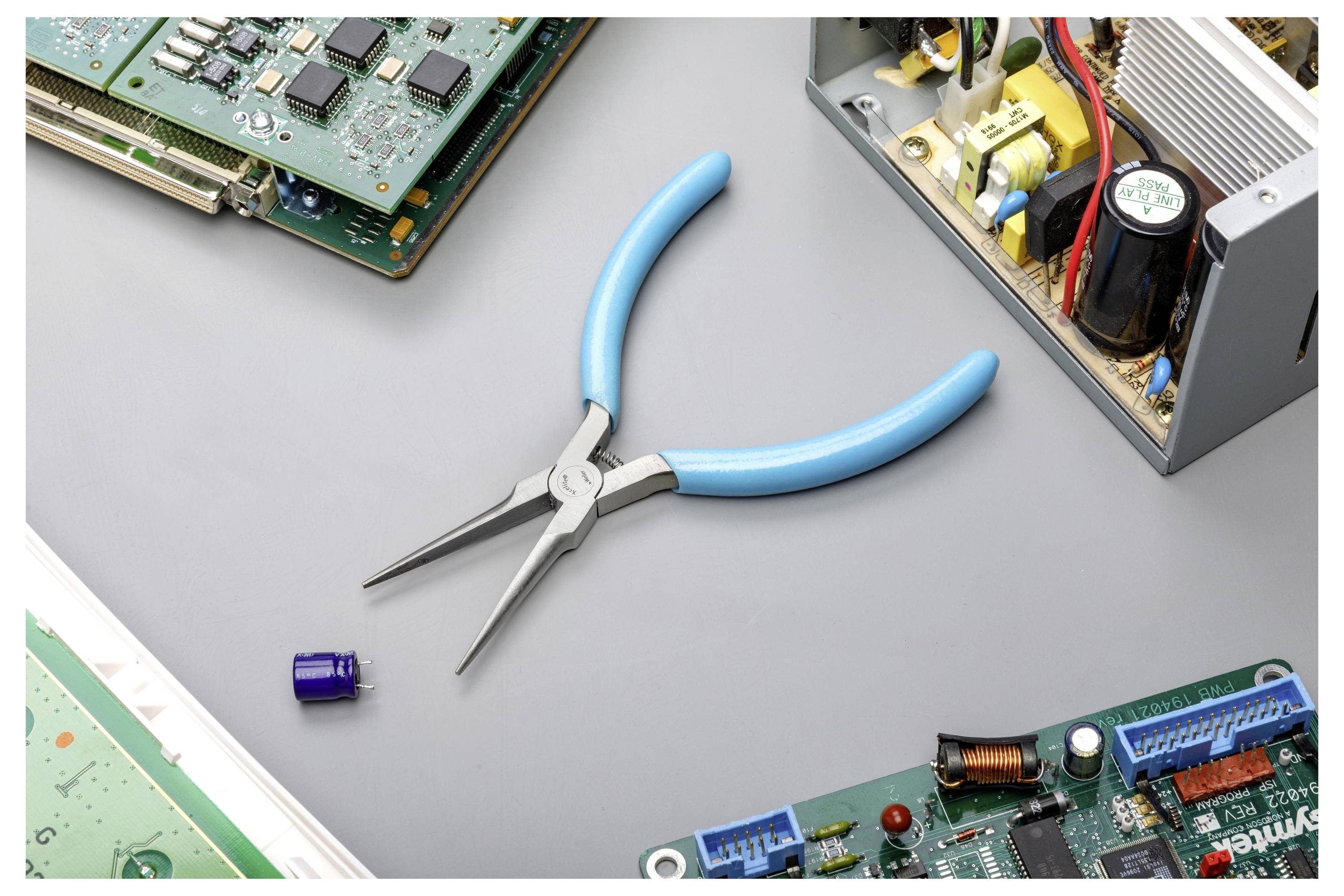 Blaugriffige Spitzzange, zentriert auf einer grauen Arbeitsfläche, umgeben von elektronischen Leiterplatten und einer geöffneten Stromversorgungskomponente.