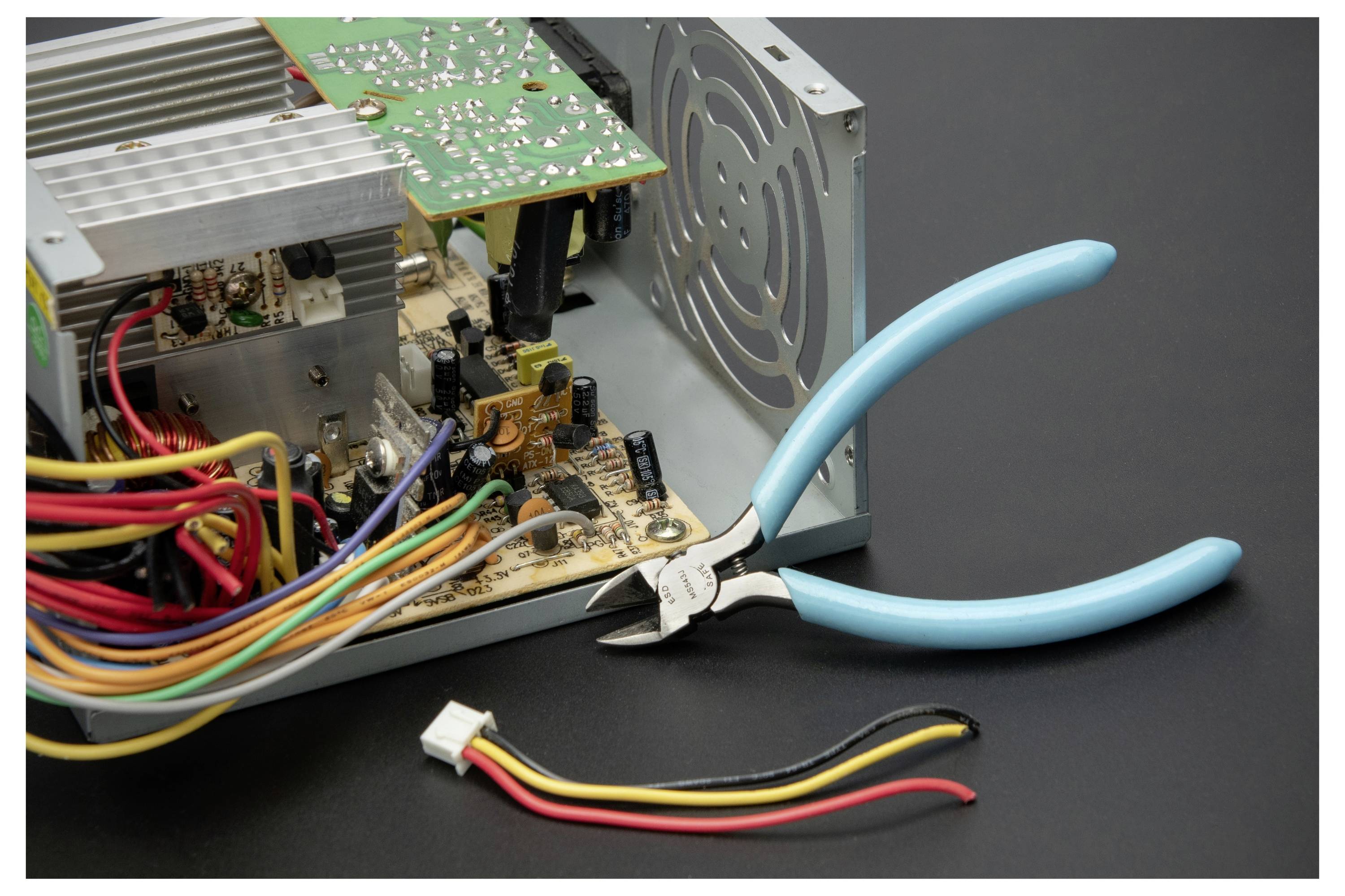 Ein Computernetzteil ist zur Reparatur geöffnet, mit freiliegenden Leiterplatten, Drähten, einem Kühlventilator und blaugriffigen Zangen daneben.