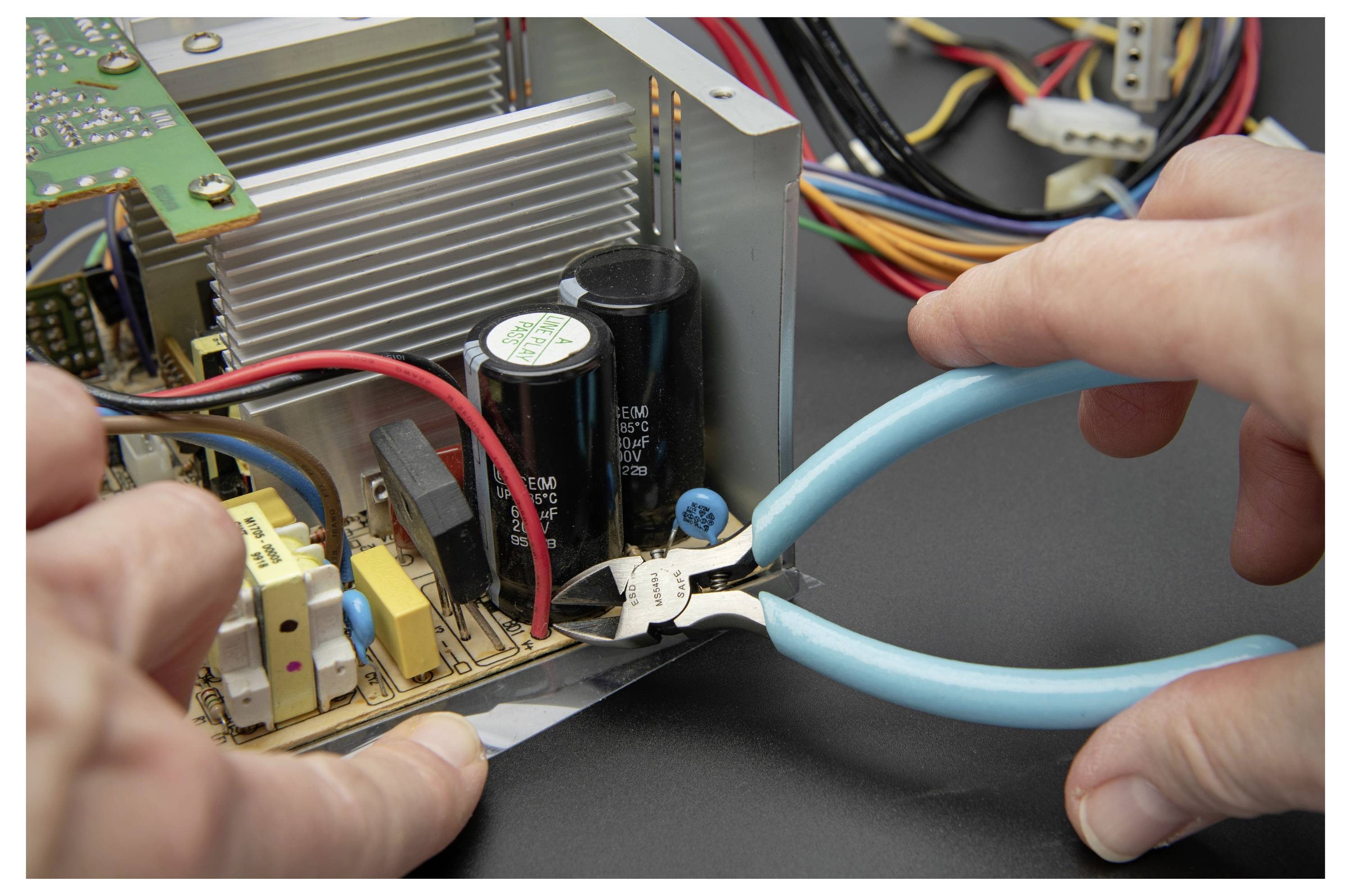 Hände, die blaugriffige Zangen verwenden, um ein weißes Kabel in einem elektronischen Gerät mit freigelegten Leiterplatten und Komponenten durchzuschneiden.